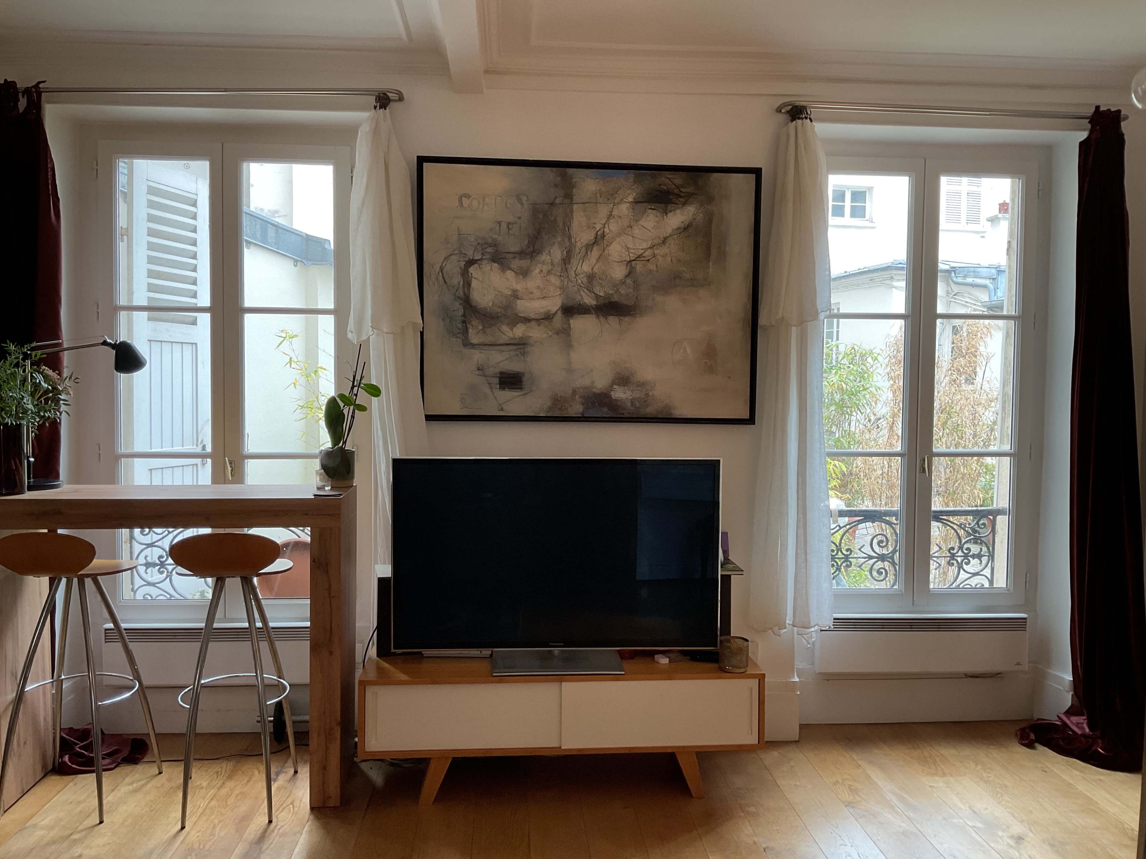 The image shows a living room with a modern TV stand, a large abstract painting on the wall, and a bar area with two stools, all framed by large windows.