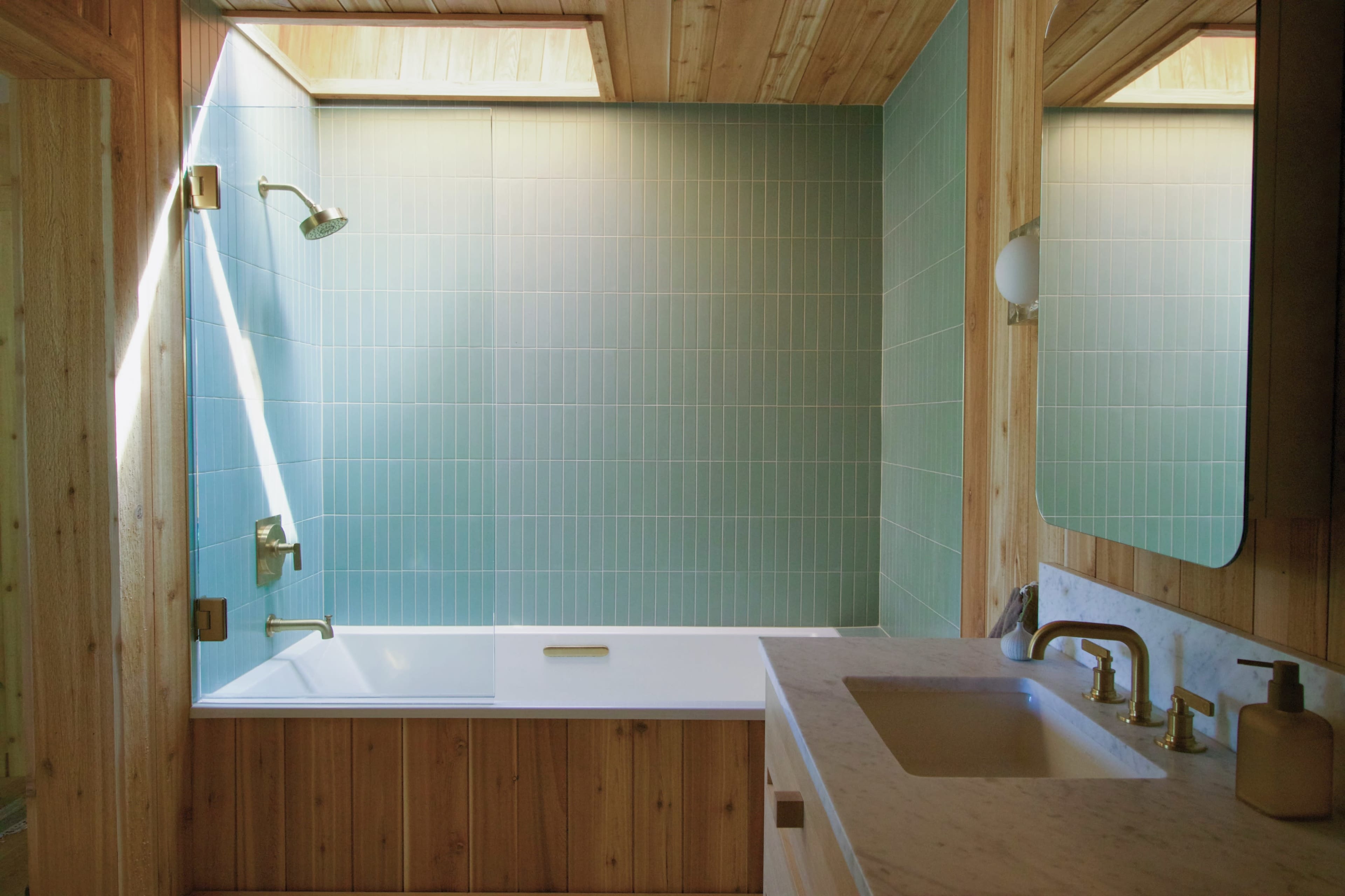 The image shows a bathroom featuring a wooden ceiling, green tiled walls, a white bathtub, and a sink with a marble countertop and gold fixtures.
