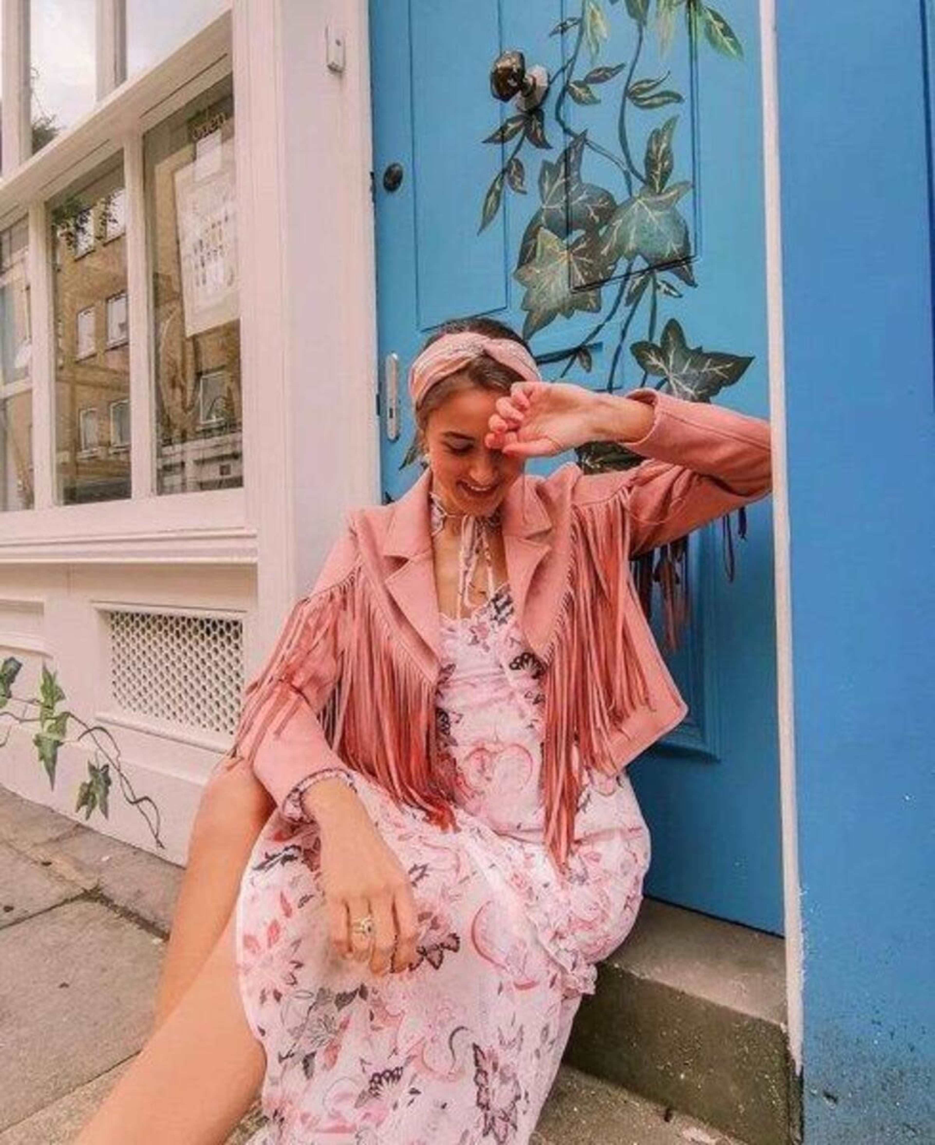 A woman in a floral dress and fringe jacket sits by a blue door adorned with a vine mural.