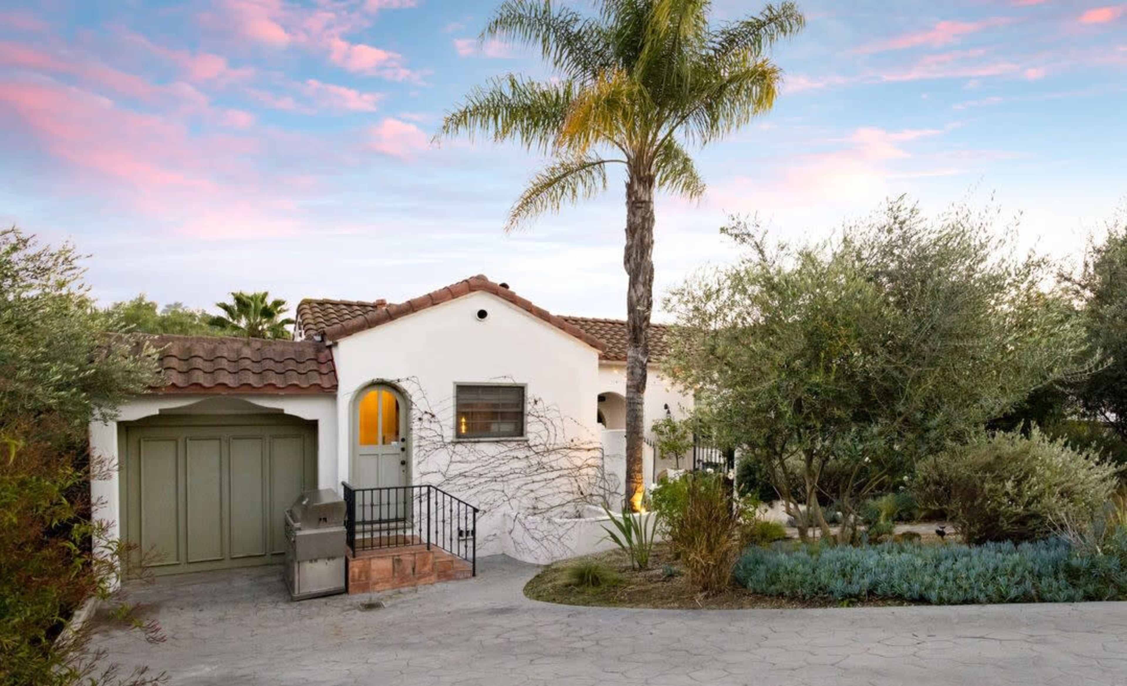 A white stucco house with a tiled roof, set amidst a landscaped garden, features a palm tree and a stone pathway leading to the entrance.