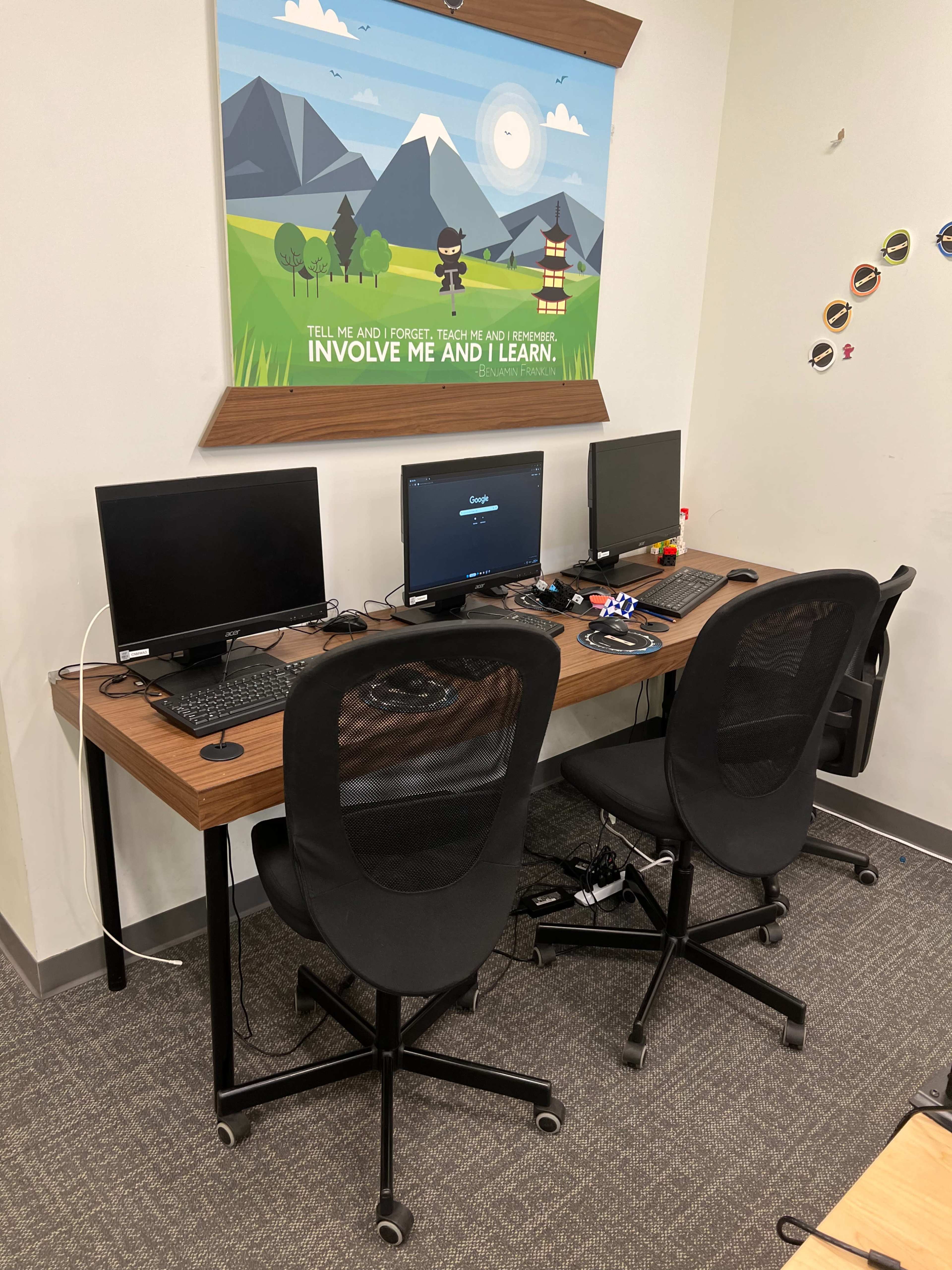 A workspace features two computer desks with monitors, located against a wall adorned with a decorative poster.