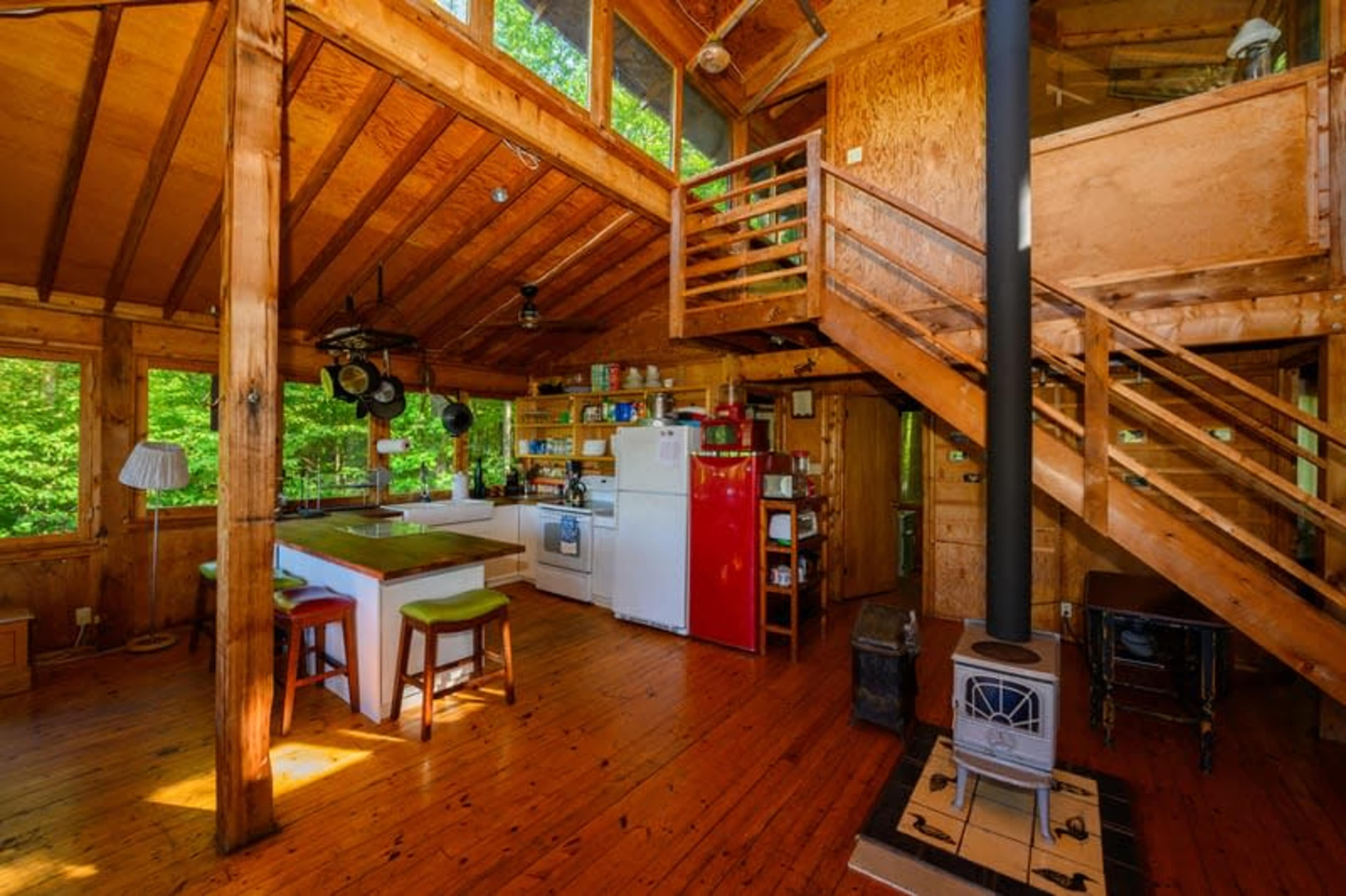 The interior of a wooden cabin with an open kitchen, a staircase, and a cozy living area featuring a stove.