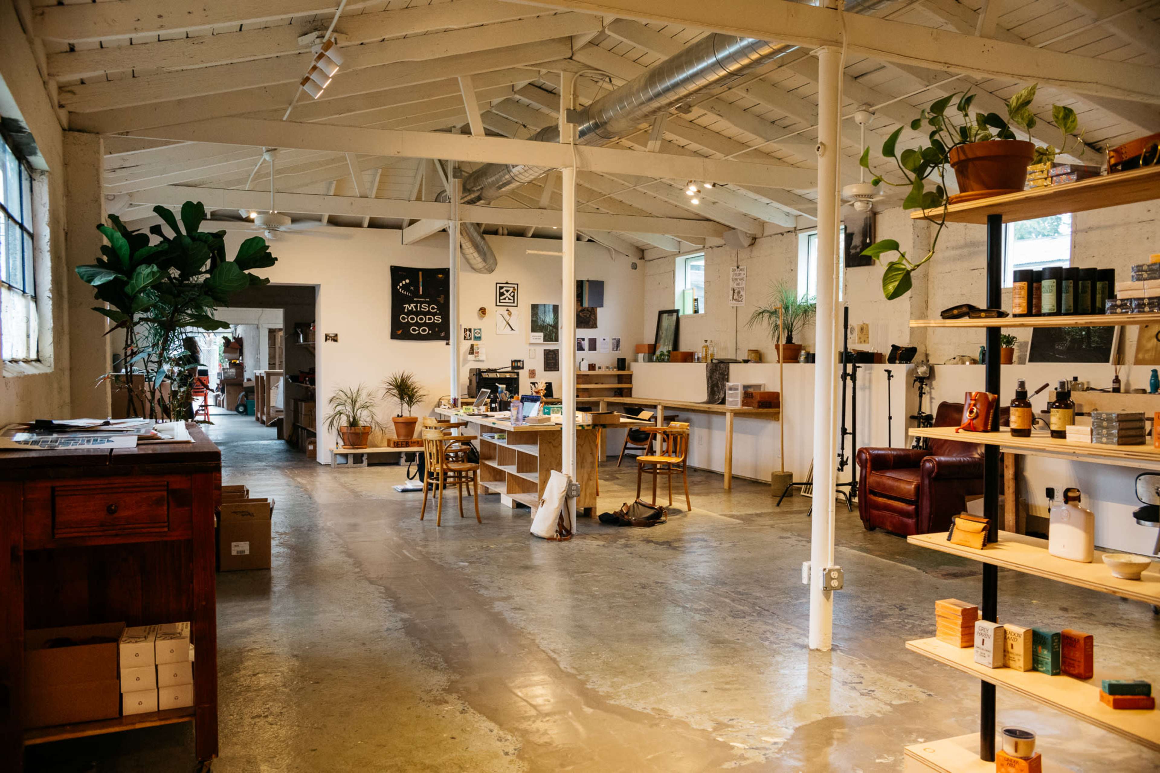 A spacious interior of a light-filled workshop features a mix of workstations, plants, and shelves displaying various products.