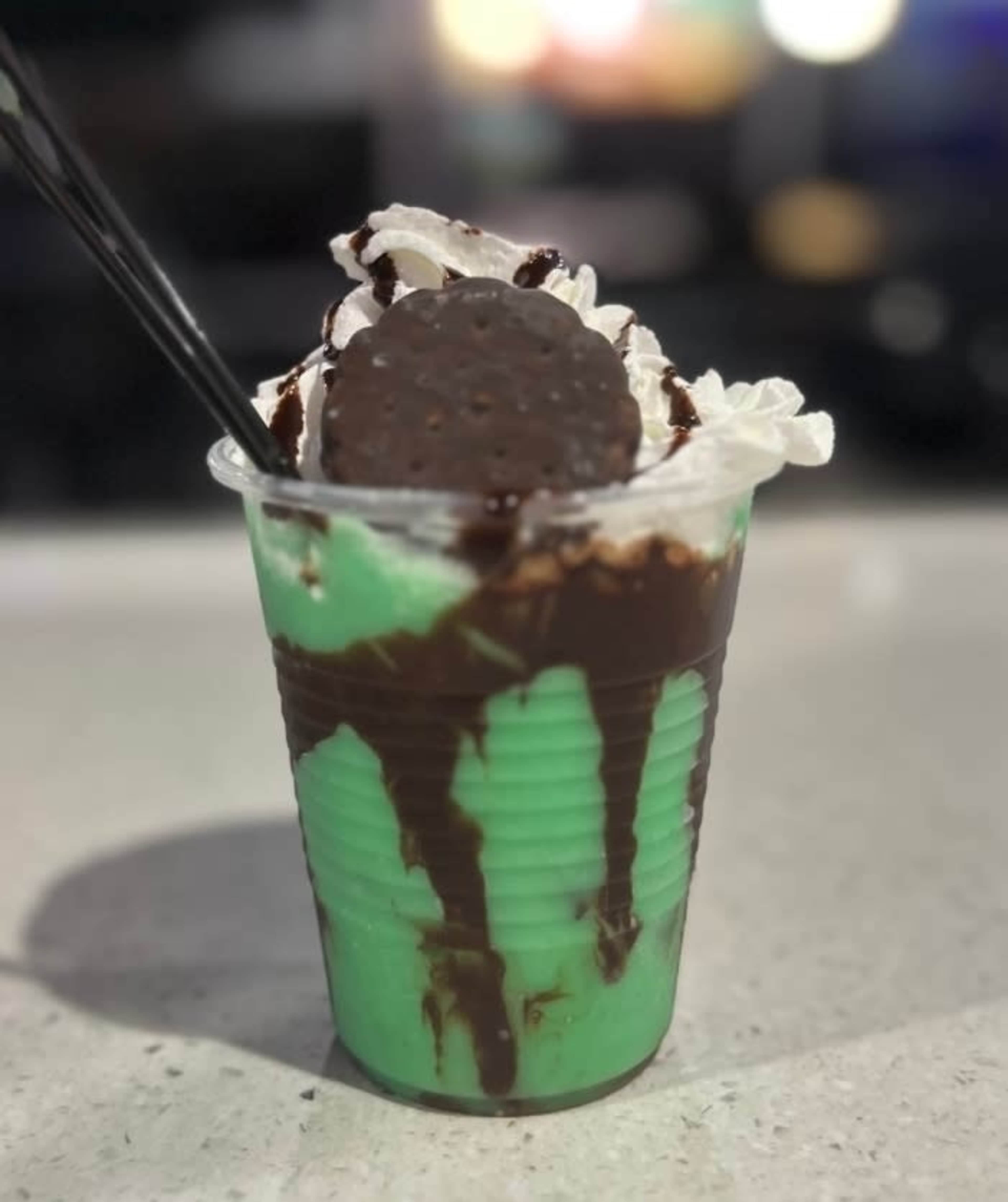 A green dessert drink topped with whipped cream, chocolate drizzle, and a chocolate wafer is placed in a clear cup with two black straws.
