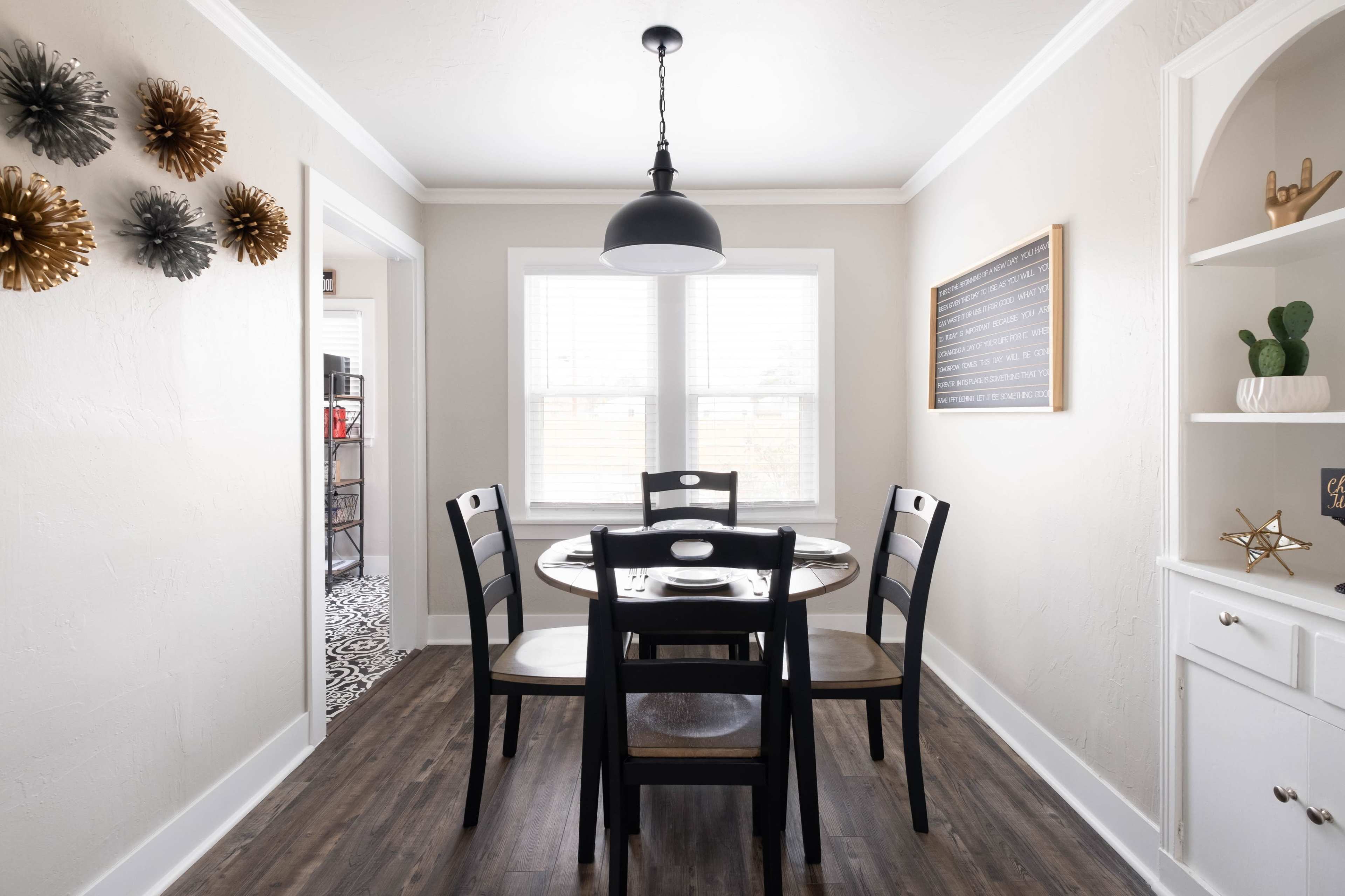A dining area features a round table with four black chairs, a black pendant light, and decorative wall elements, alongside a white cabinet and a chalkboard.