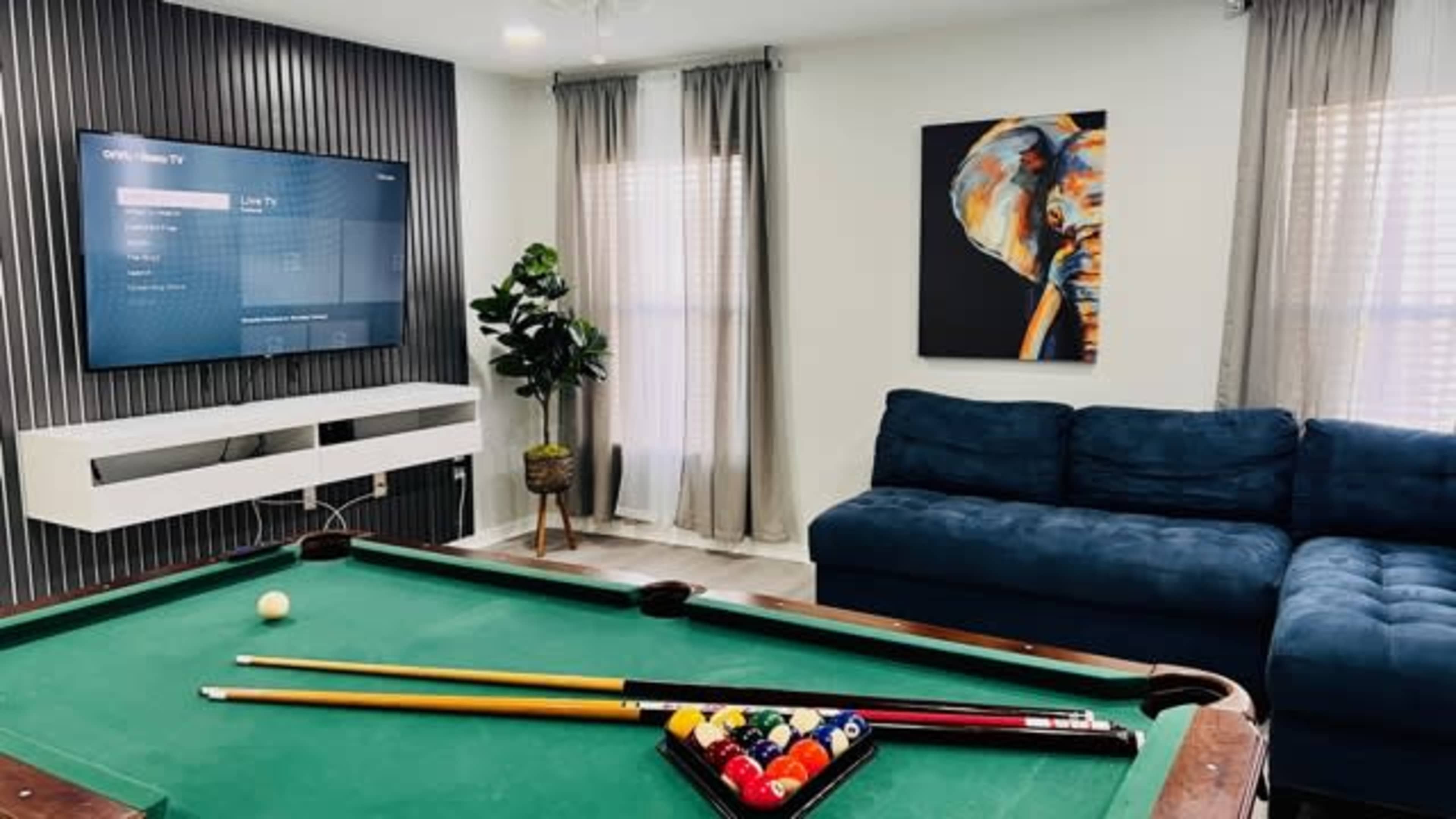 A living room featuring a pool table with colorful balls, a blue couch, a television mounted on the wall, and a framed painting of a figure.