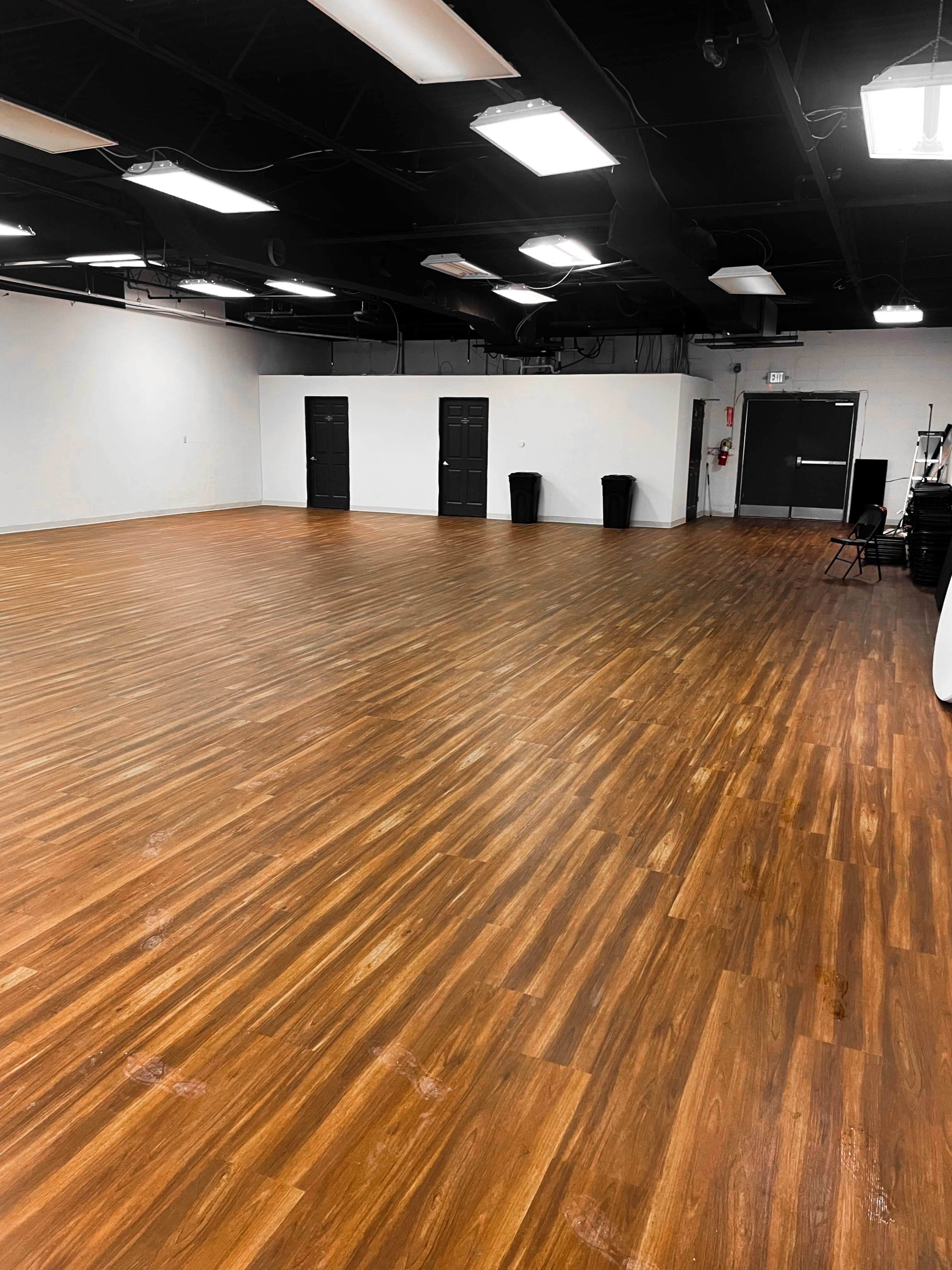 The image shows a spacious, empty room with wooden floors and black doors along one wall.