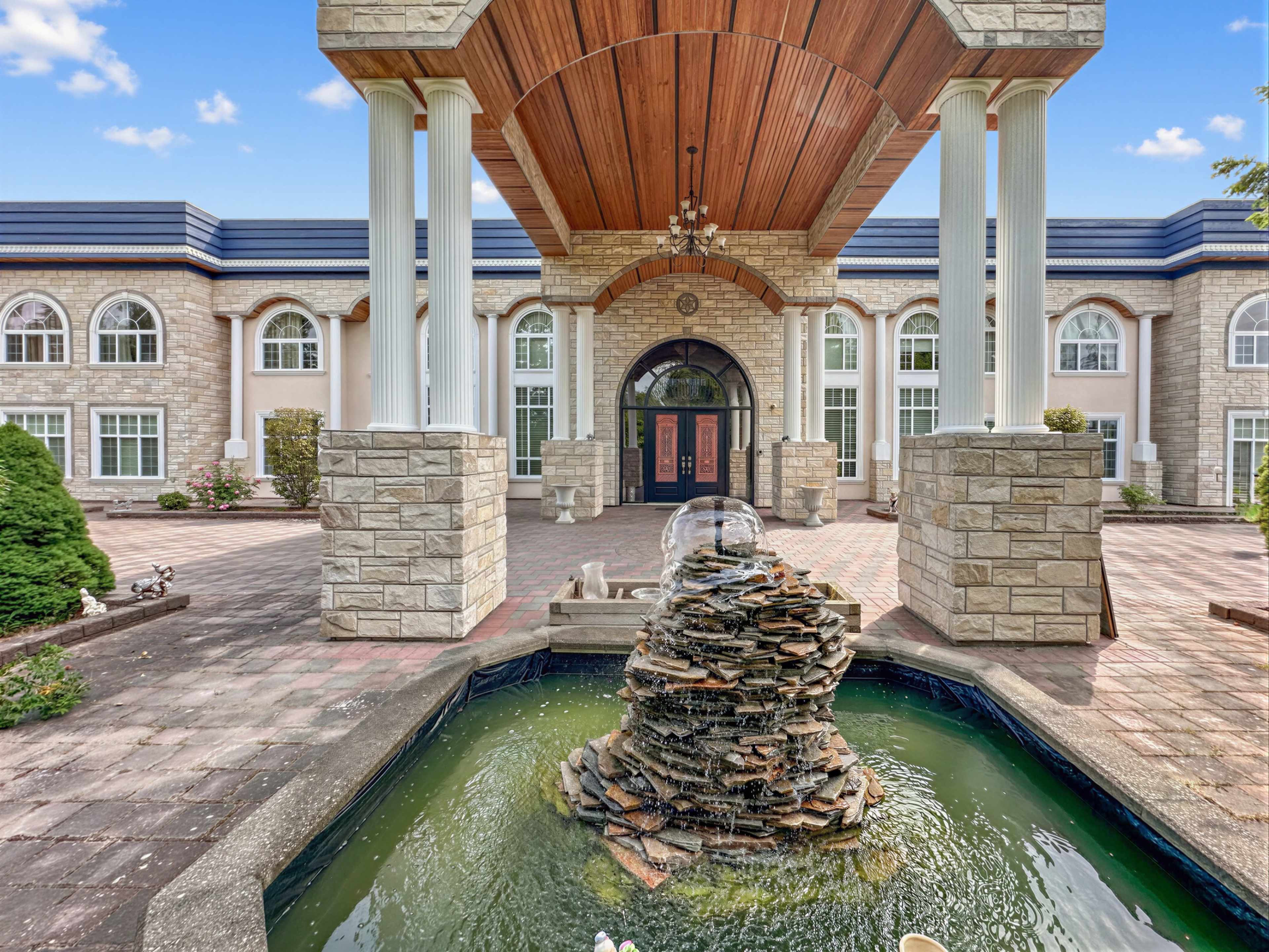 The image shows a grand entrance of a building with stone columns, a wooden ceiling, and a central fountain surrounded by greenery and paved walkways.