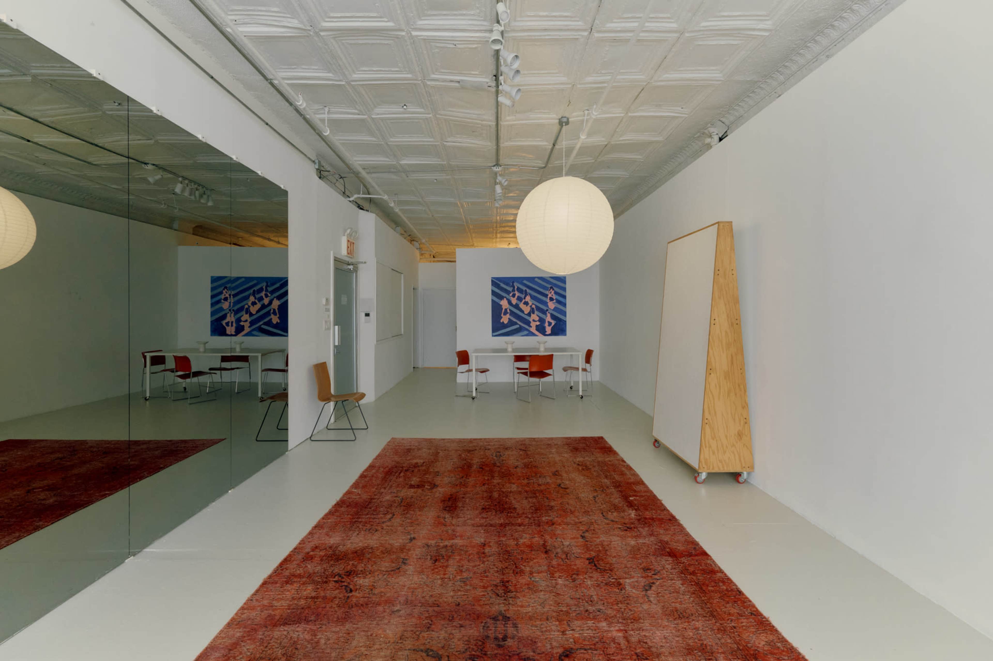 The image shows a spacious, minimalist room featuring a large rug, a table with chairs, and a large mirror on one wall.