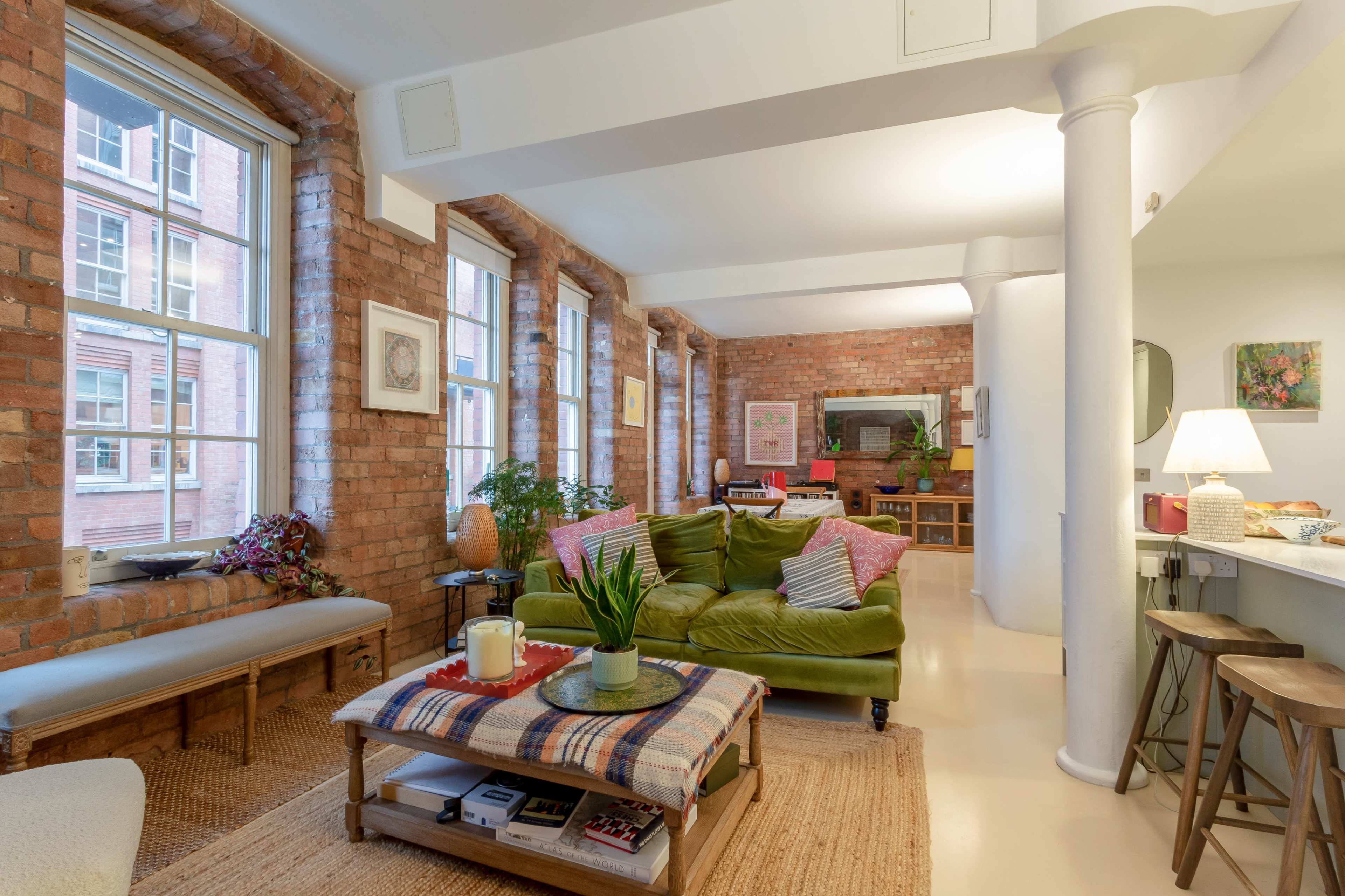 A modern living space with large windows, exposed brick walls, a green sofa, and a wooden coffee table adorned with a plant and a candle.