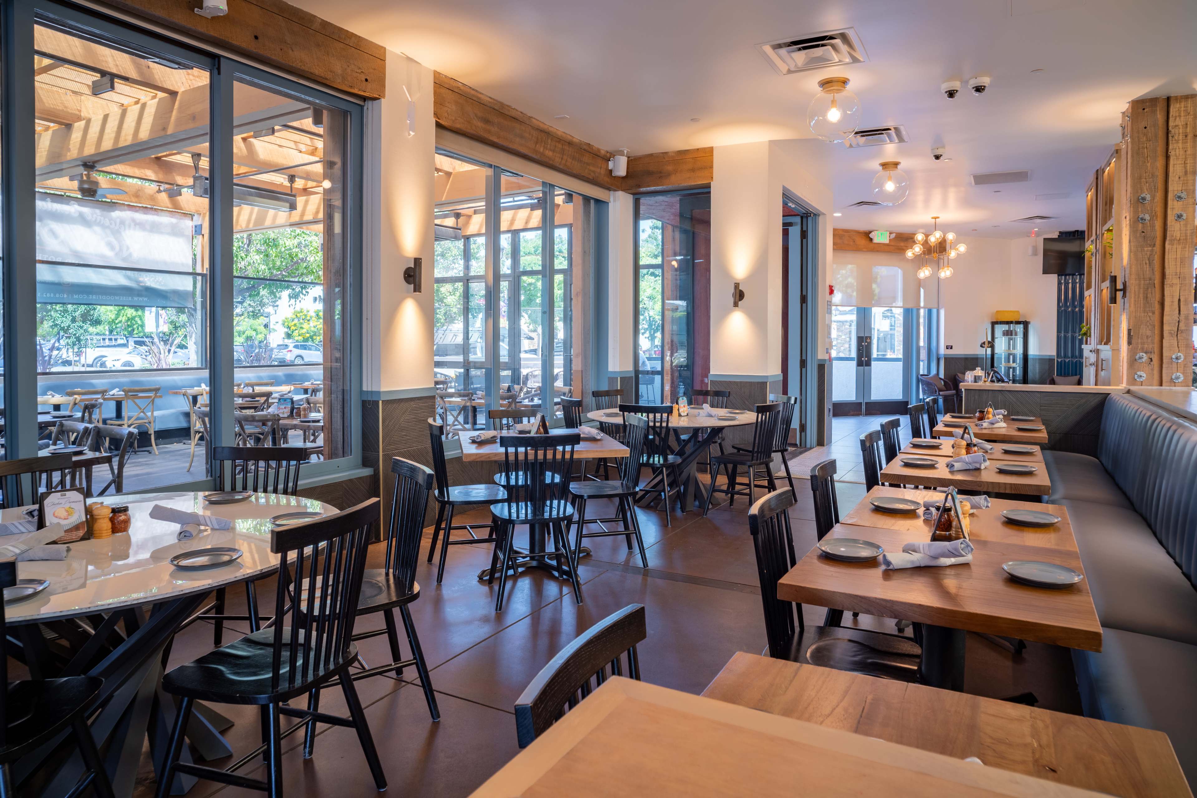 The image shows a modern restaurant interior with wooden tables, black chairs, and large windows allowing natural light to fill the space.
