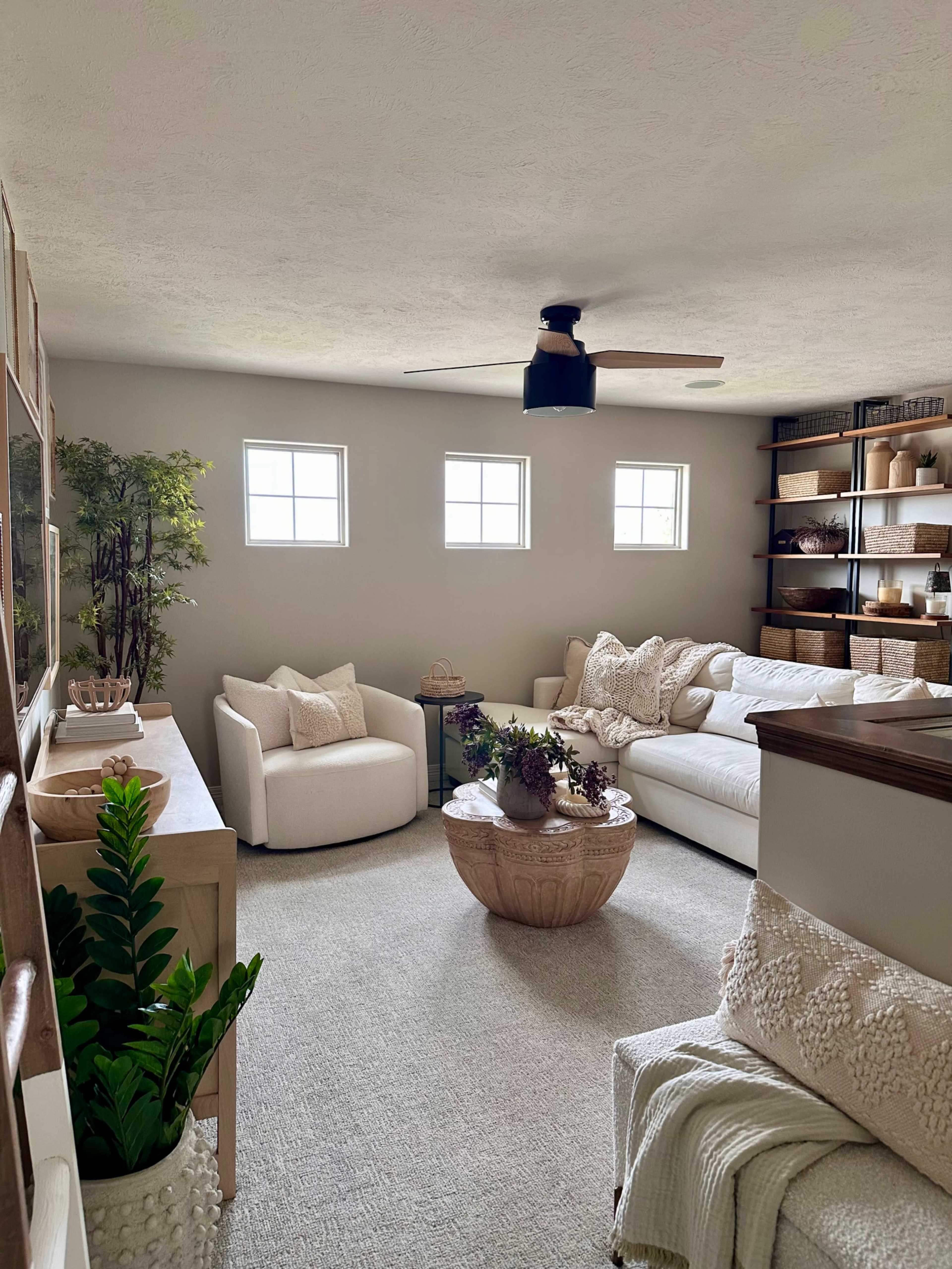 The image shows a living room with a white couch, armchair, round wooden coffee table, and shelves filled with decorative items and plants.