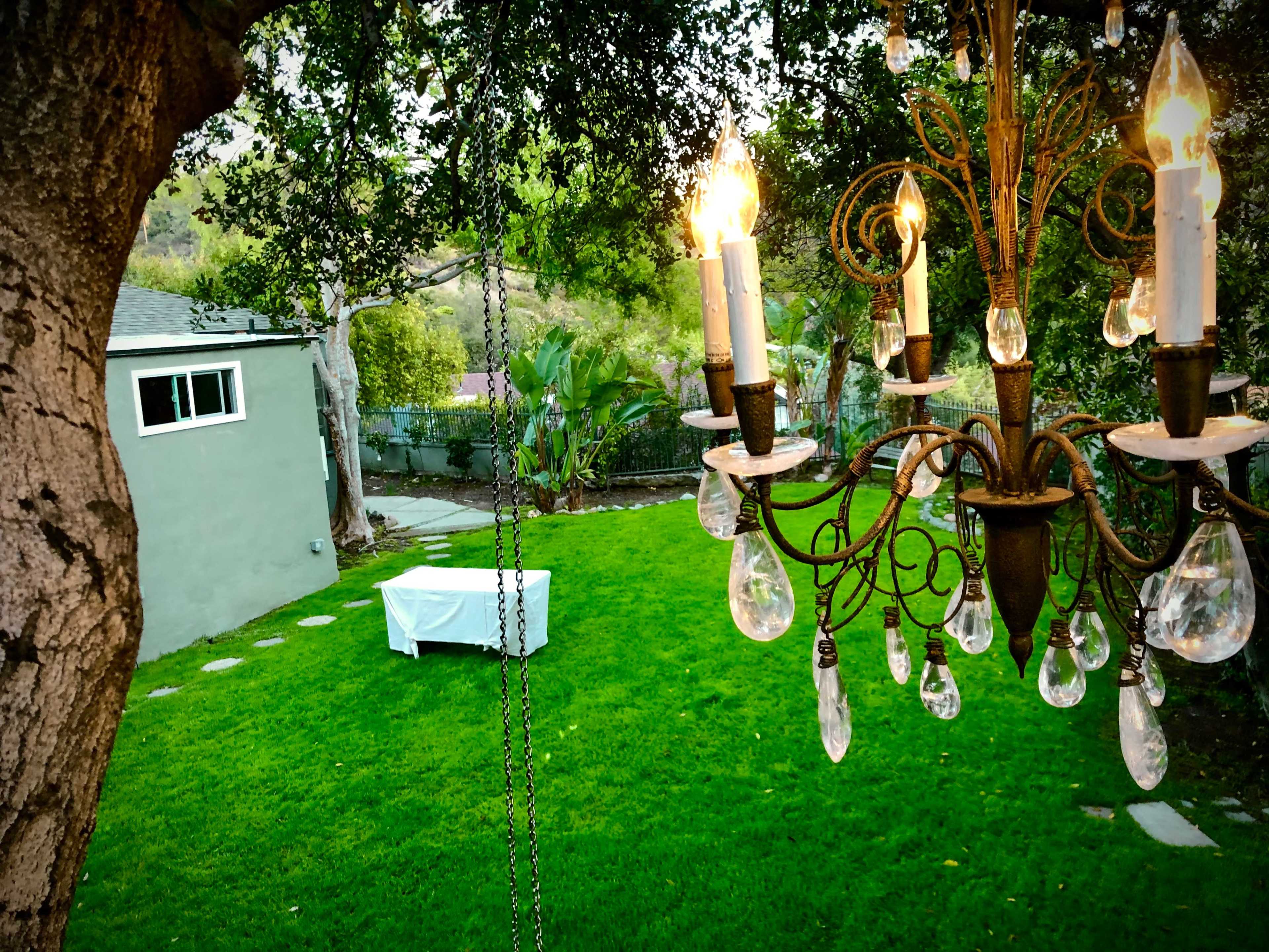 A chandelier hangs above a backyard with a neatly trimmed lawn and a table covered with a white cloth in the distance.