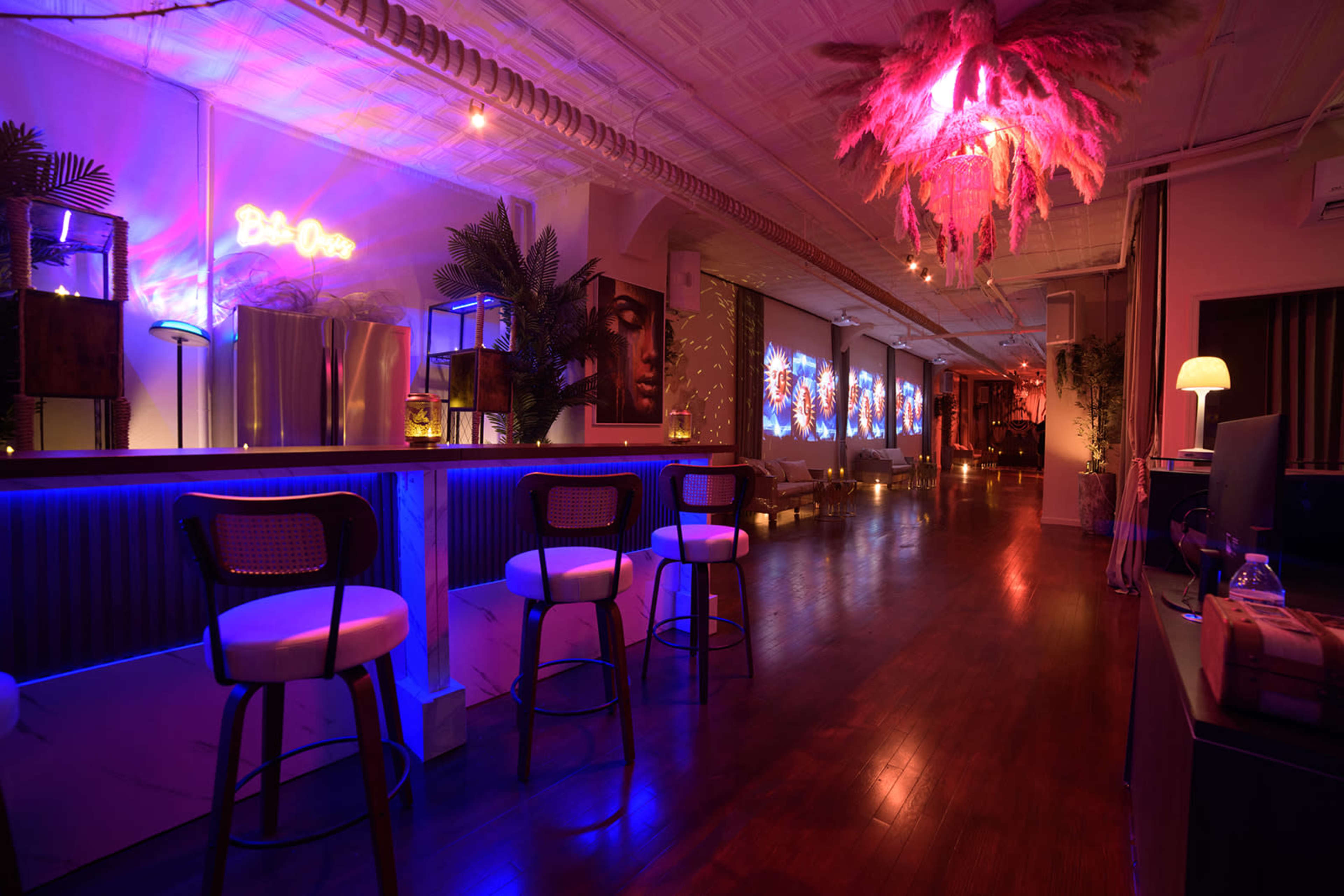 The image shows a dimly lit bar area with neon lights, modern furniture, and colorful artwork on the walls.