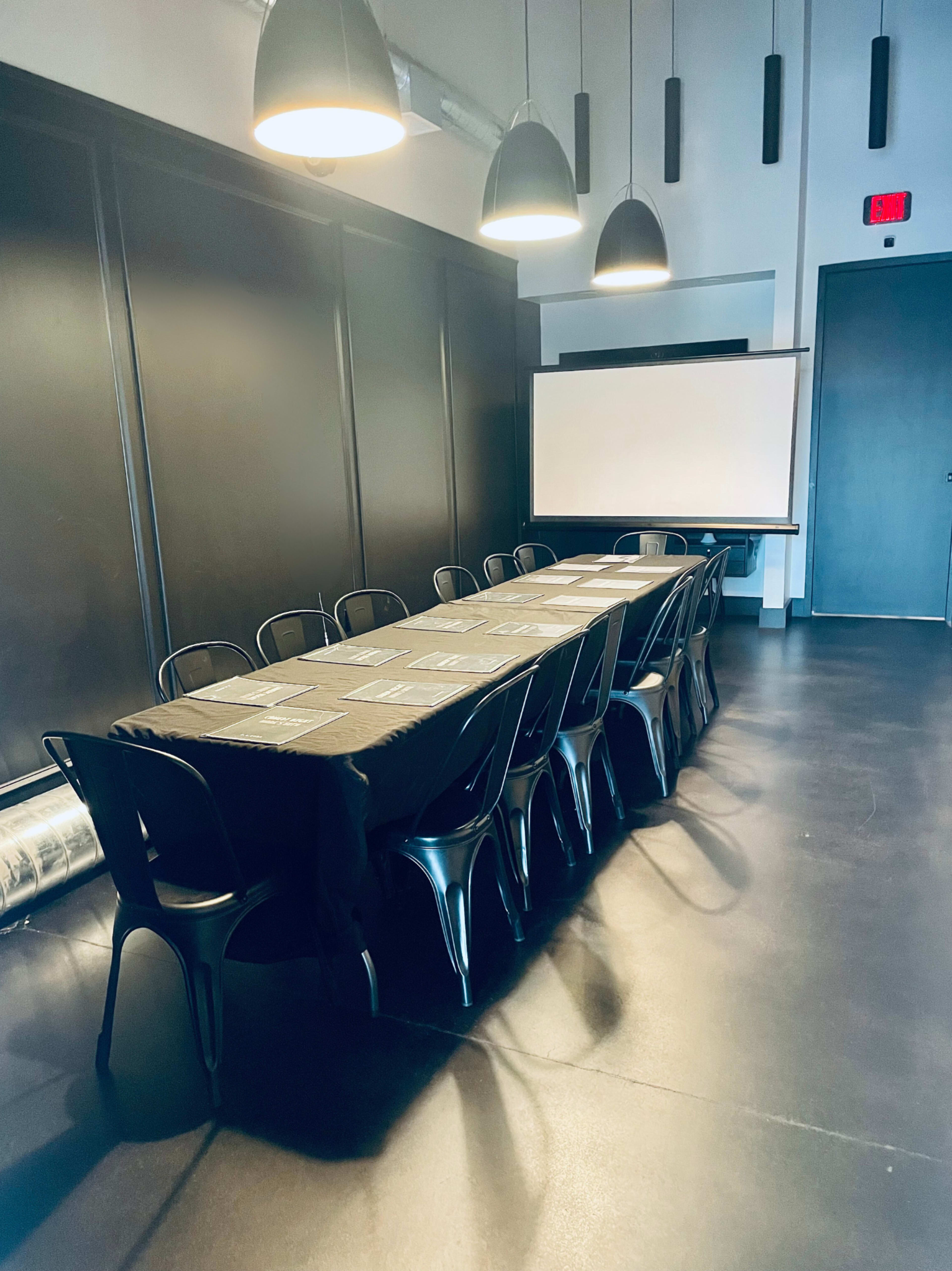 A long rectangular table with black chairs is set up in a dimly lit conference room, featuring a projector screen at one end.