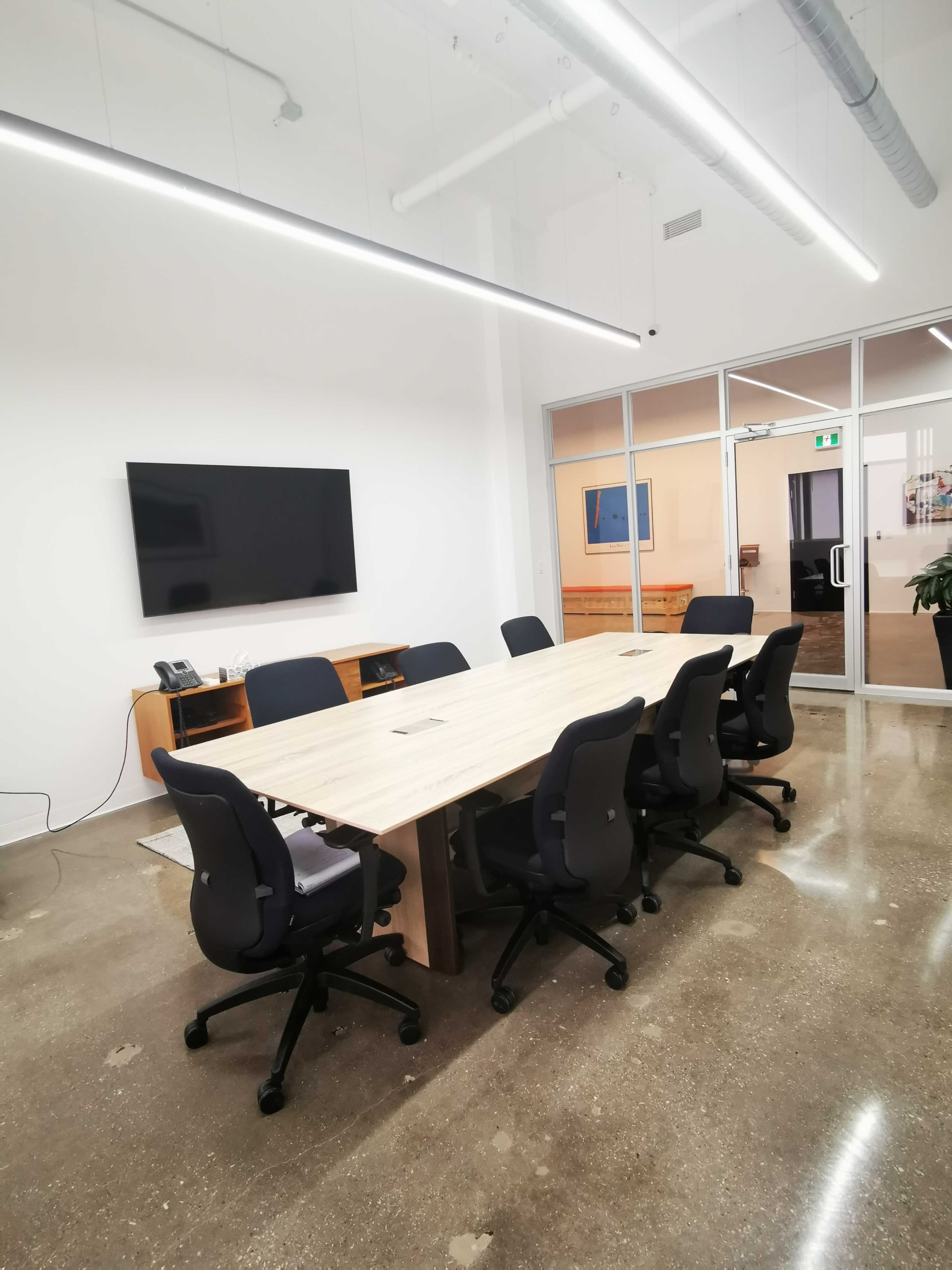 The image shows a modern conference room with a large wooden table, eight black office chairs, a wall-mounted television, and glass doors leading to another area.