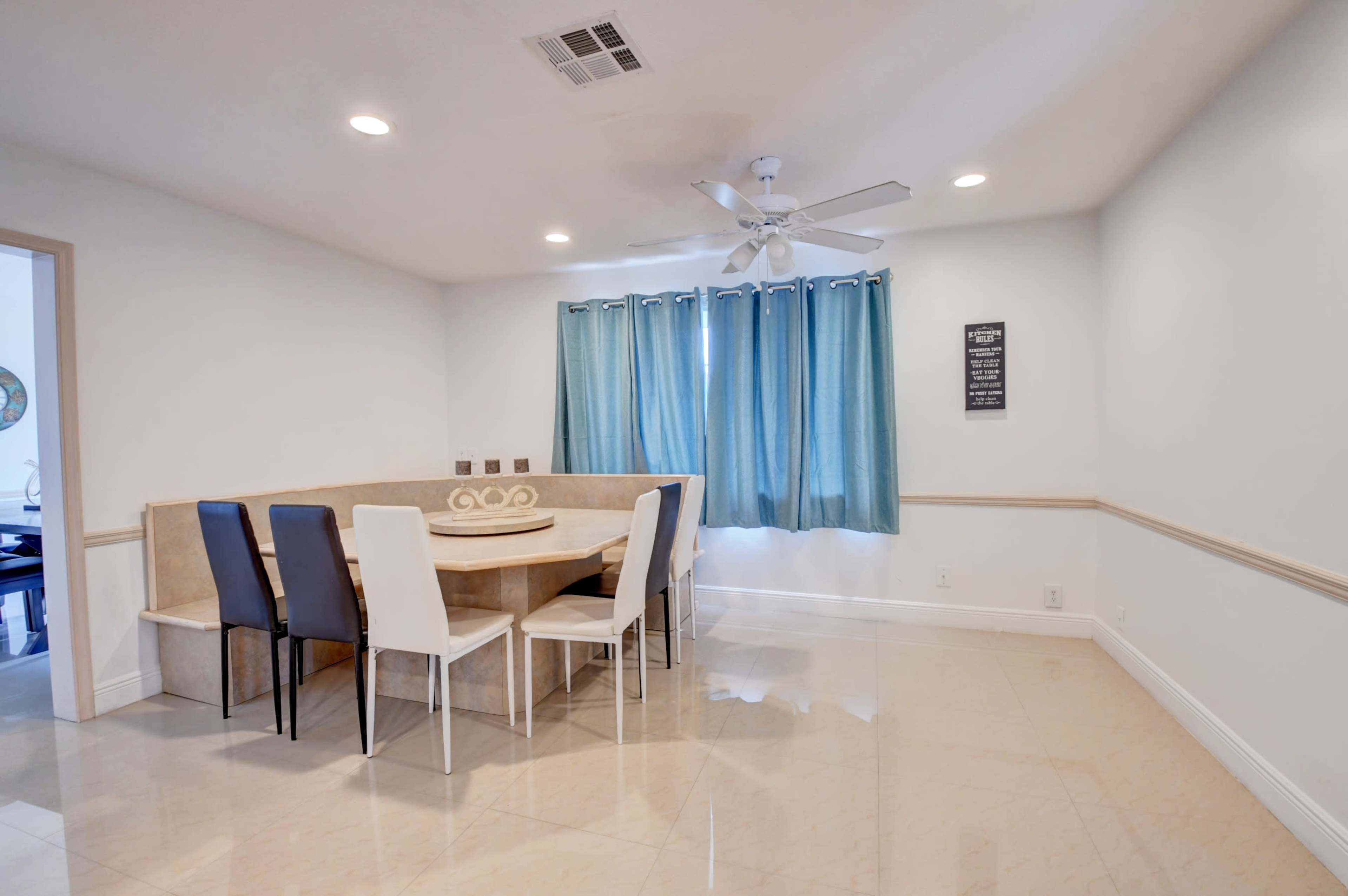 A dining area features a round table surrounded by chairs, with a ceiling fan and large windows dressed in turquoise curtains.