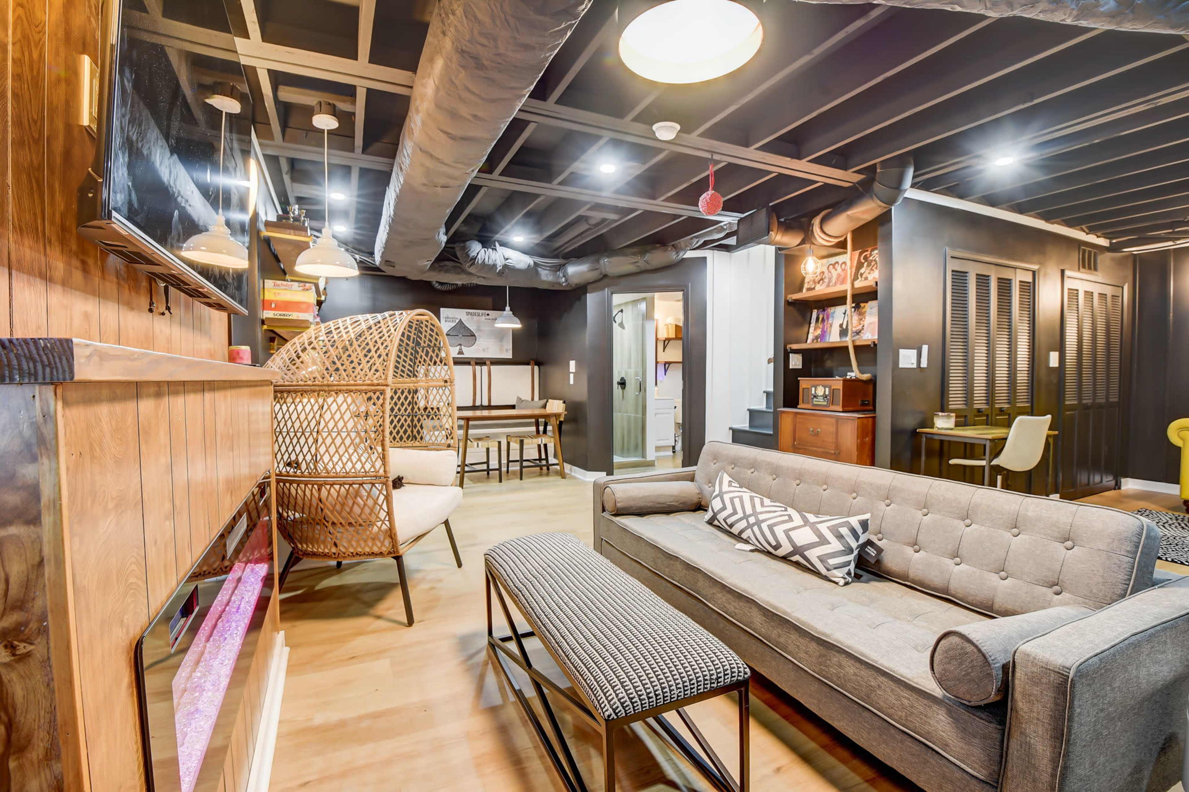 The image shows a modern basement living room with a sofa, a coffee table, a rattan chair, and a kitchenette area in the background.