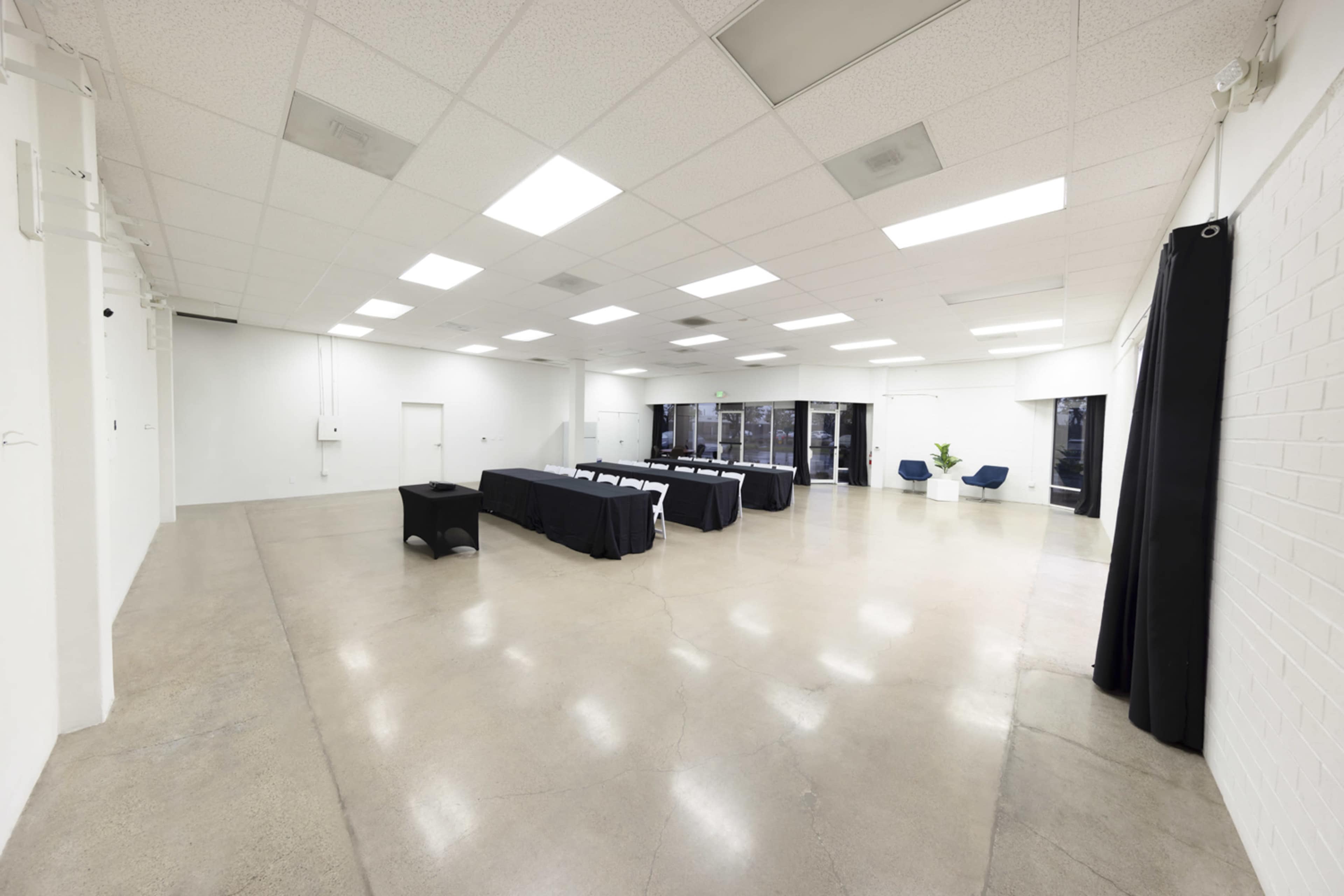 An empty, spacious room with polished concrete floors, white walls, and a long table set with black tablecloths at one end.
