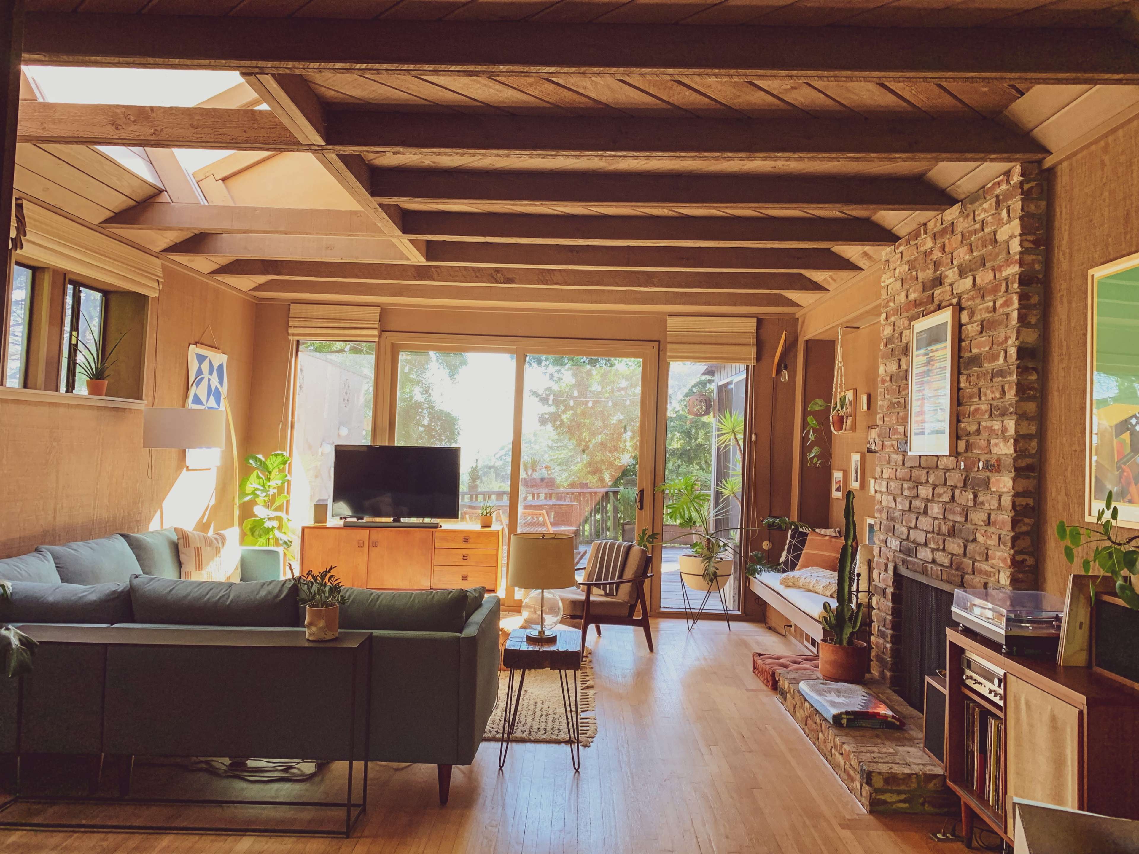 A cozy living room with wooden beams, a large window overlooking greenery, a sectional sofa, a television, and a stone fireplace.