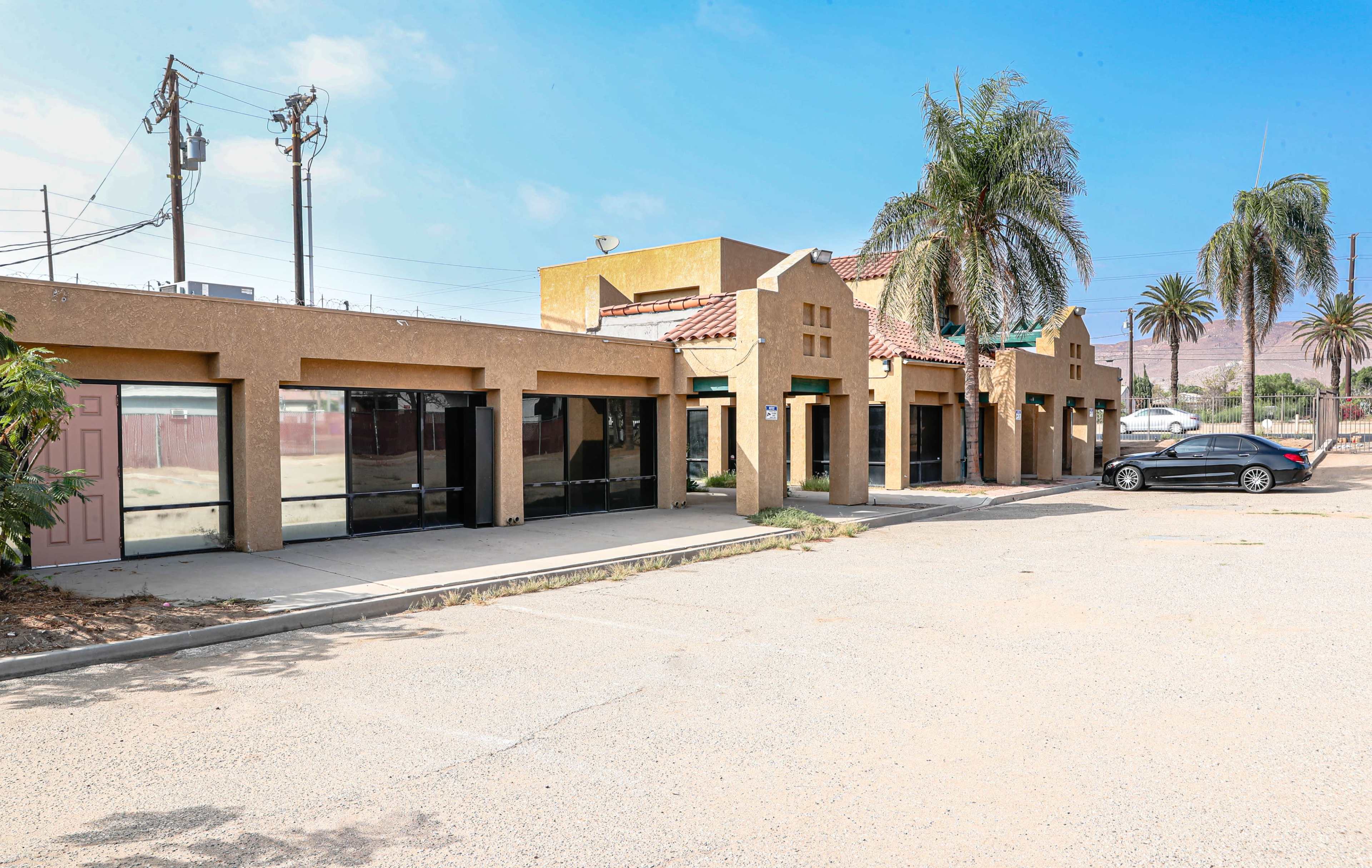 A row of single-story commercial buildings with a stucco facade and tile roofs sits adjacent to a parking lot lined with palm trees.