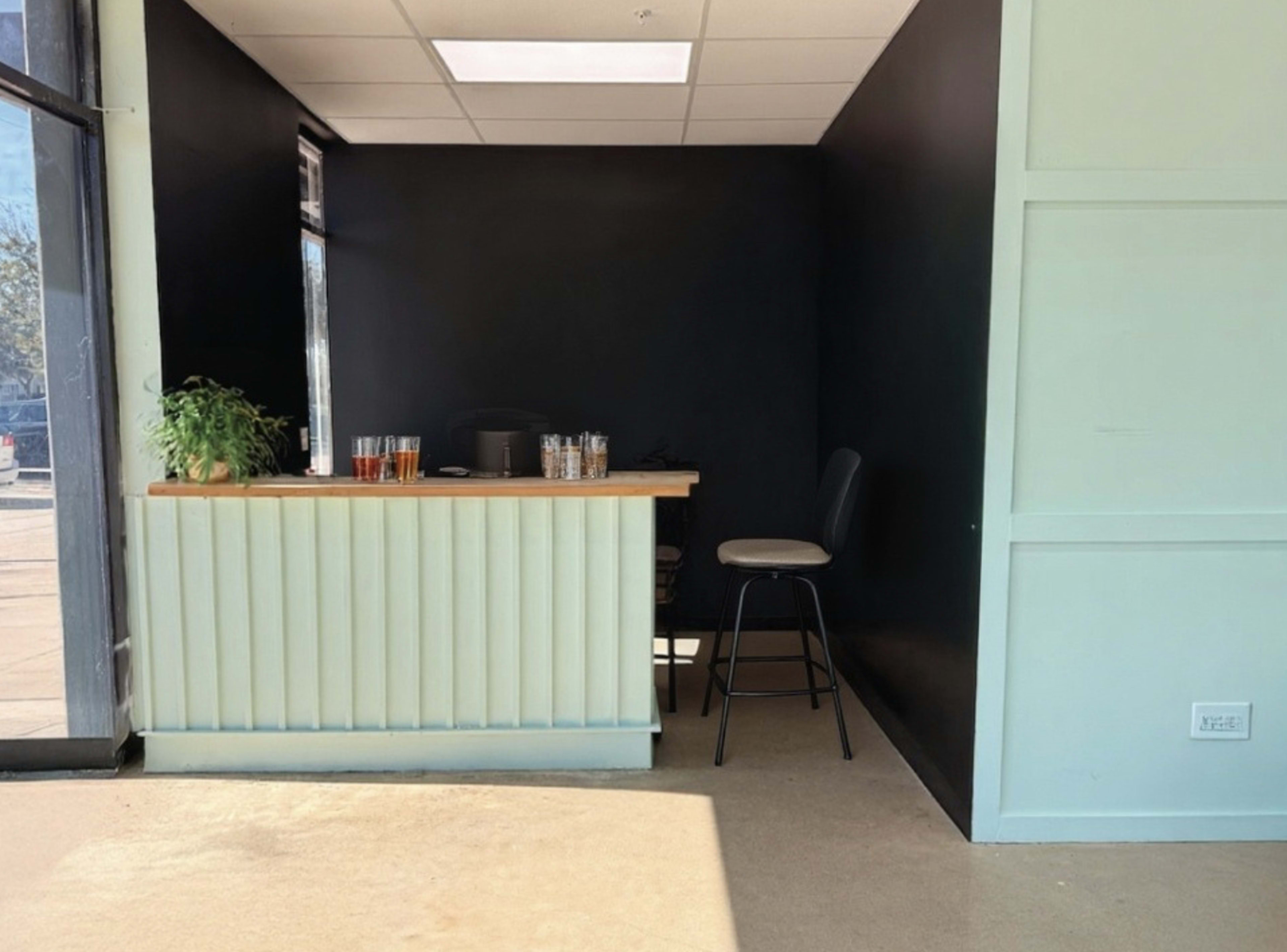 The image shows a small, minimalistic reception area with a light-colored counter, several glasses on the surface, and a single barstool against a dark wall.