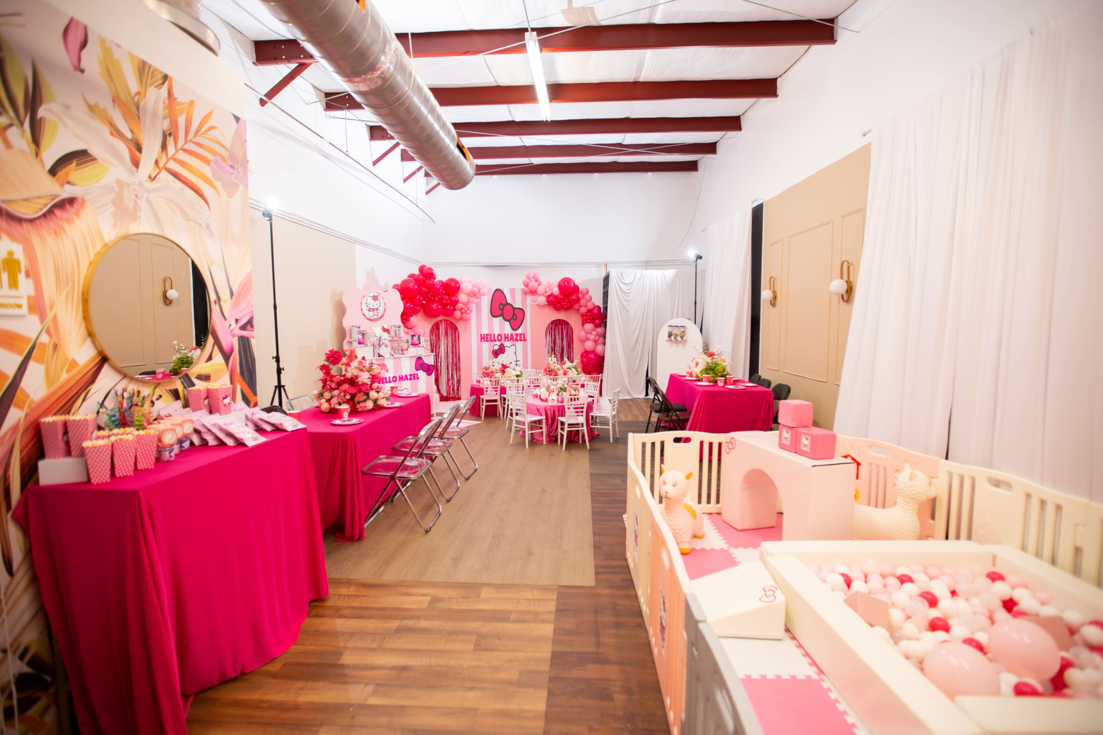 The image shows a decorated party venue featuring pink tables, balloons, and a play area with a ball pit.
