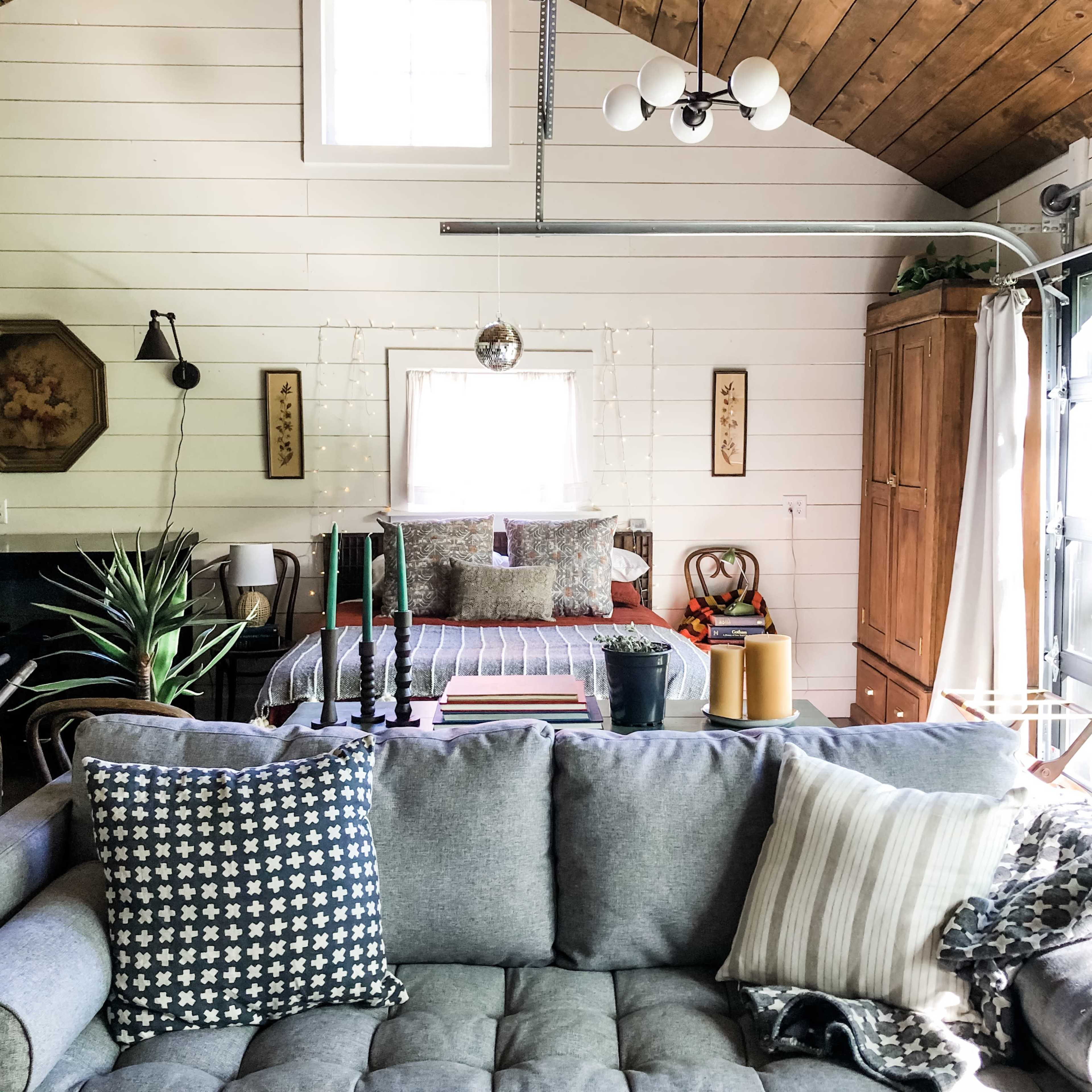 A cozy living space features a gray couch adorned with decorative pillows, a bed in the background, and wooden accents on the walls.