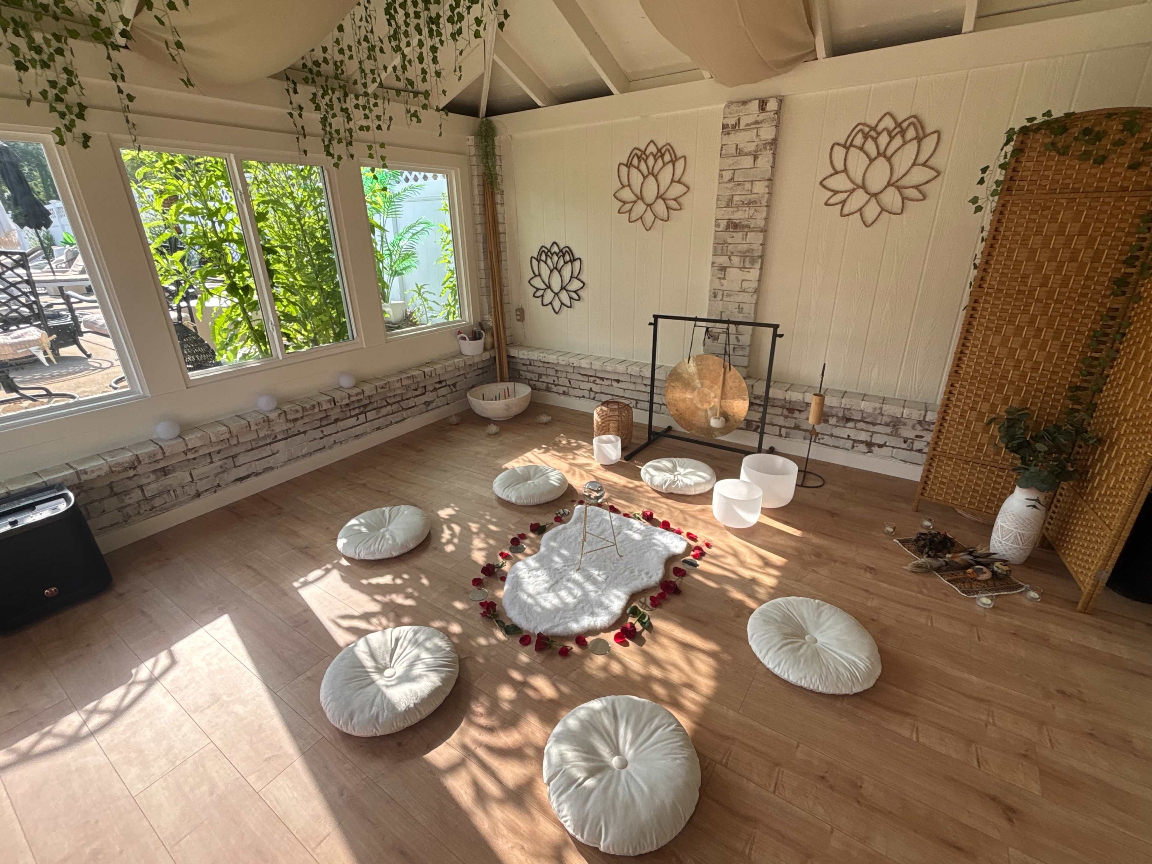 The image shows a serene meditation space with a circular arrangement of floor cushions, a gong, and various decorative elements surrounded by plants and large windows.