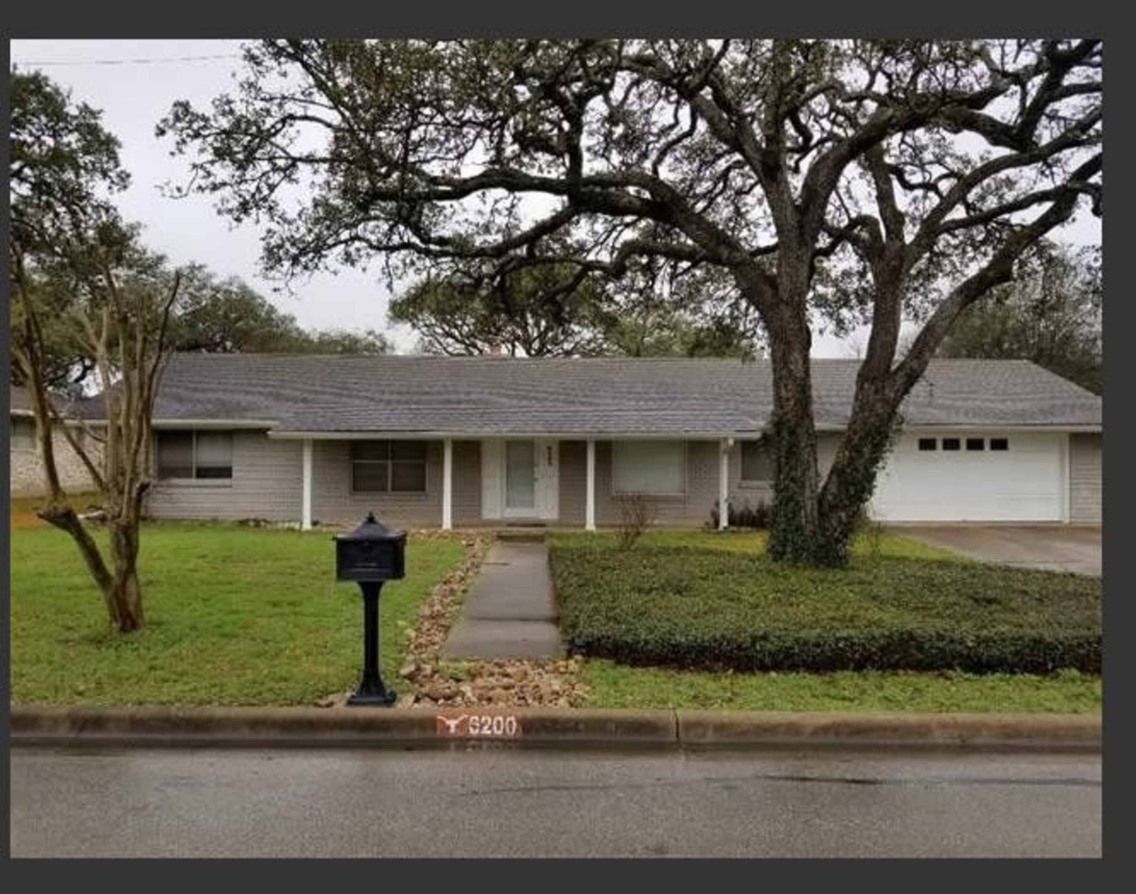 A single-story house with a gray shingled roof, a green lawn, and a large tree in the front yard.