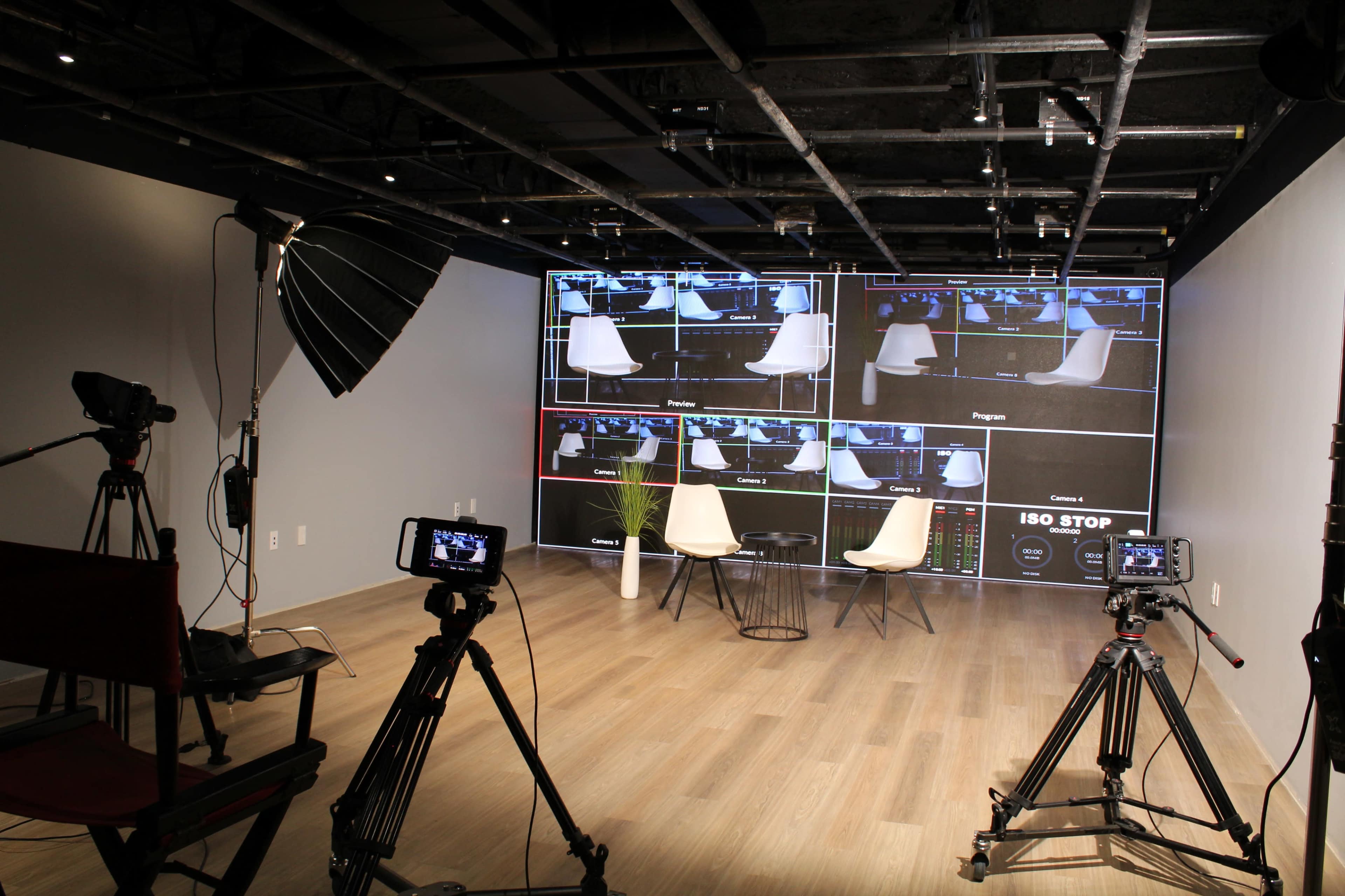 The image shows a video production studio with two empty chairs in front of multiple monitors displaying different camera feeds and equipment set up for filming.