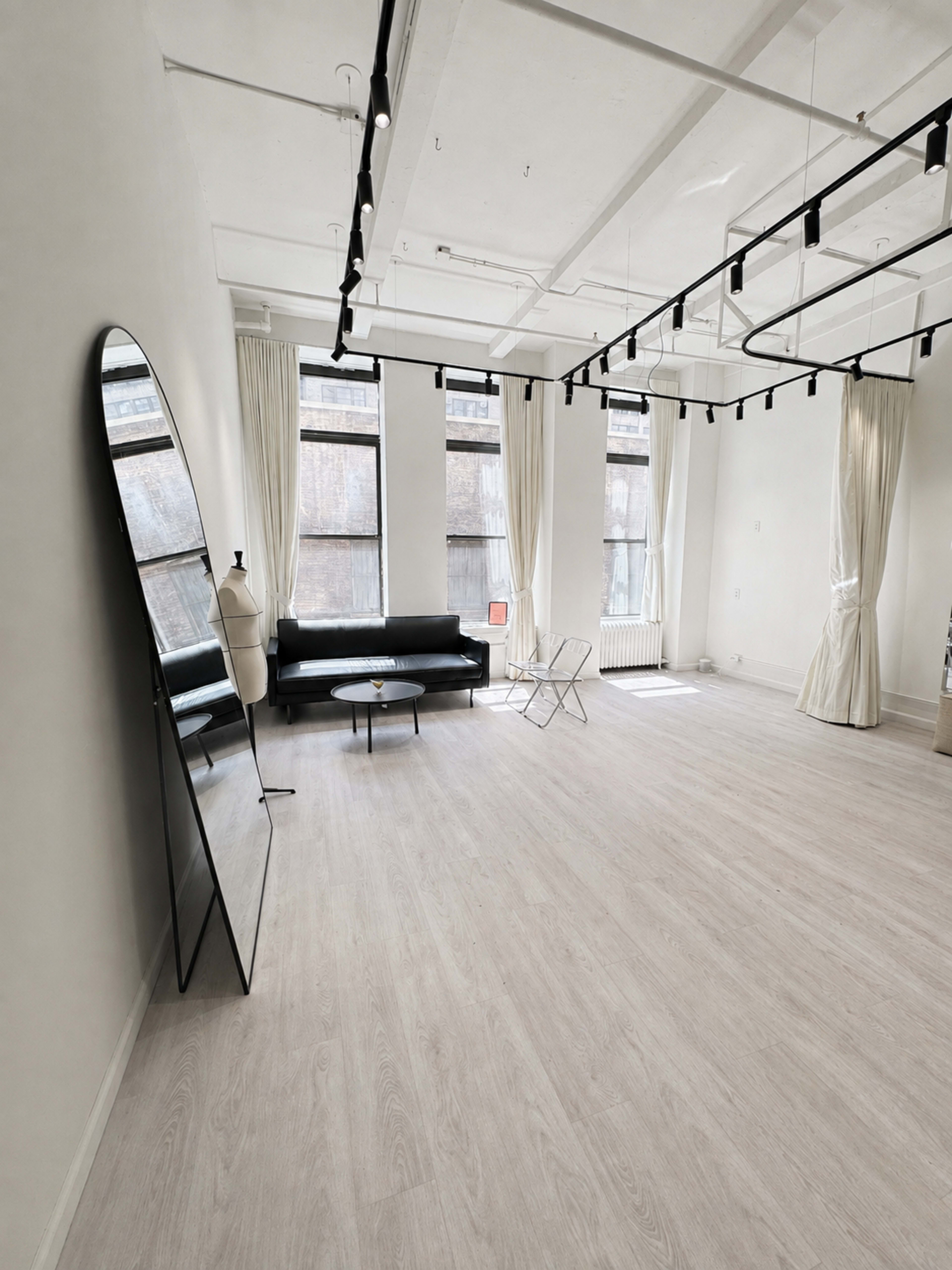 The image shows a bright, minimalist room with wooden flooring, featuring a black sofa, a mirror, and large windows with light curtains.