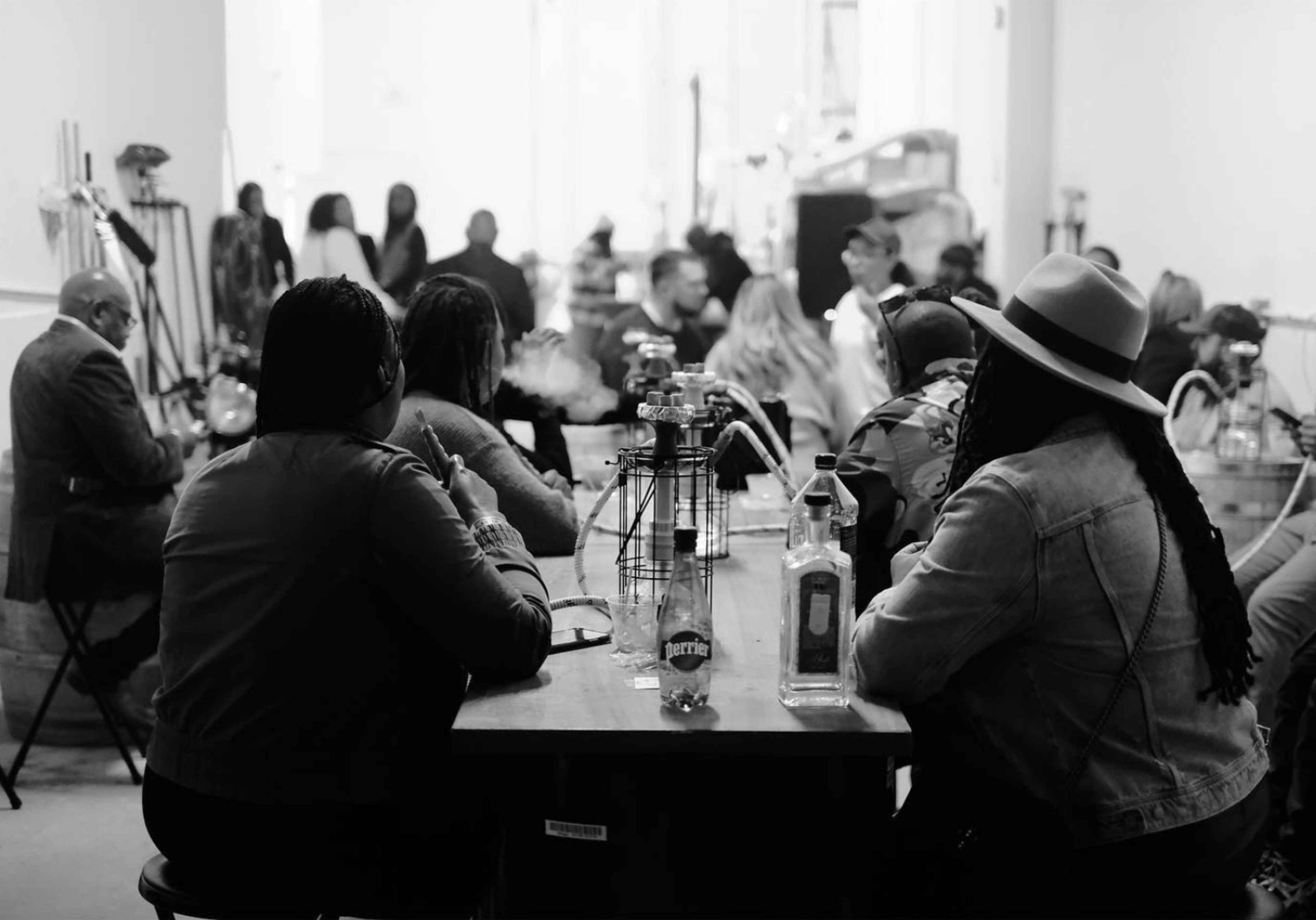 A group of people sits at a large table in a dimly lit room, with bottles and hookahs placed on the table.