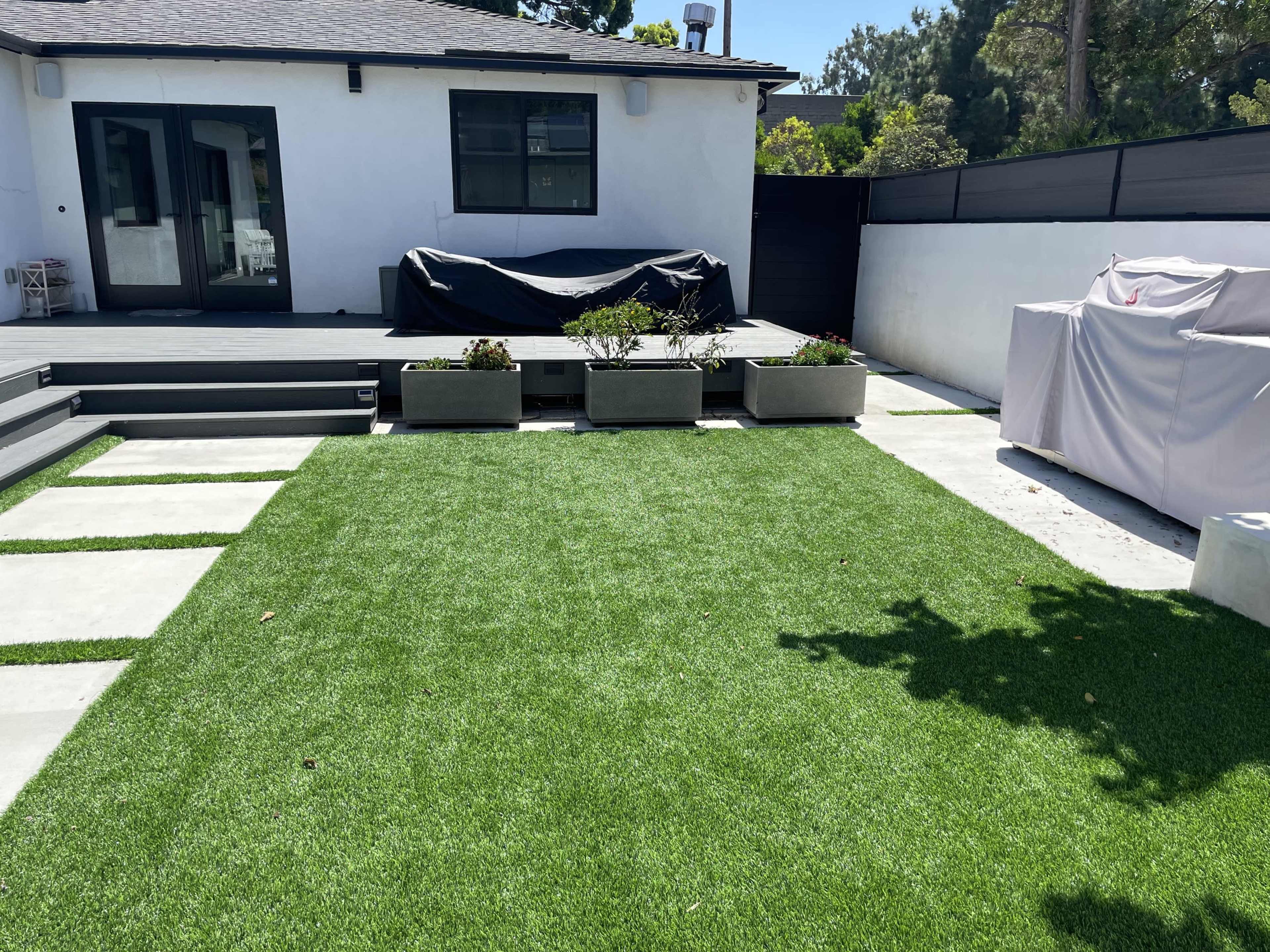 A backyard with synthetic grass, concrete pavers, and several planters in front of a white house with a deck and covered outdoor furniture.
