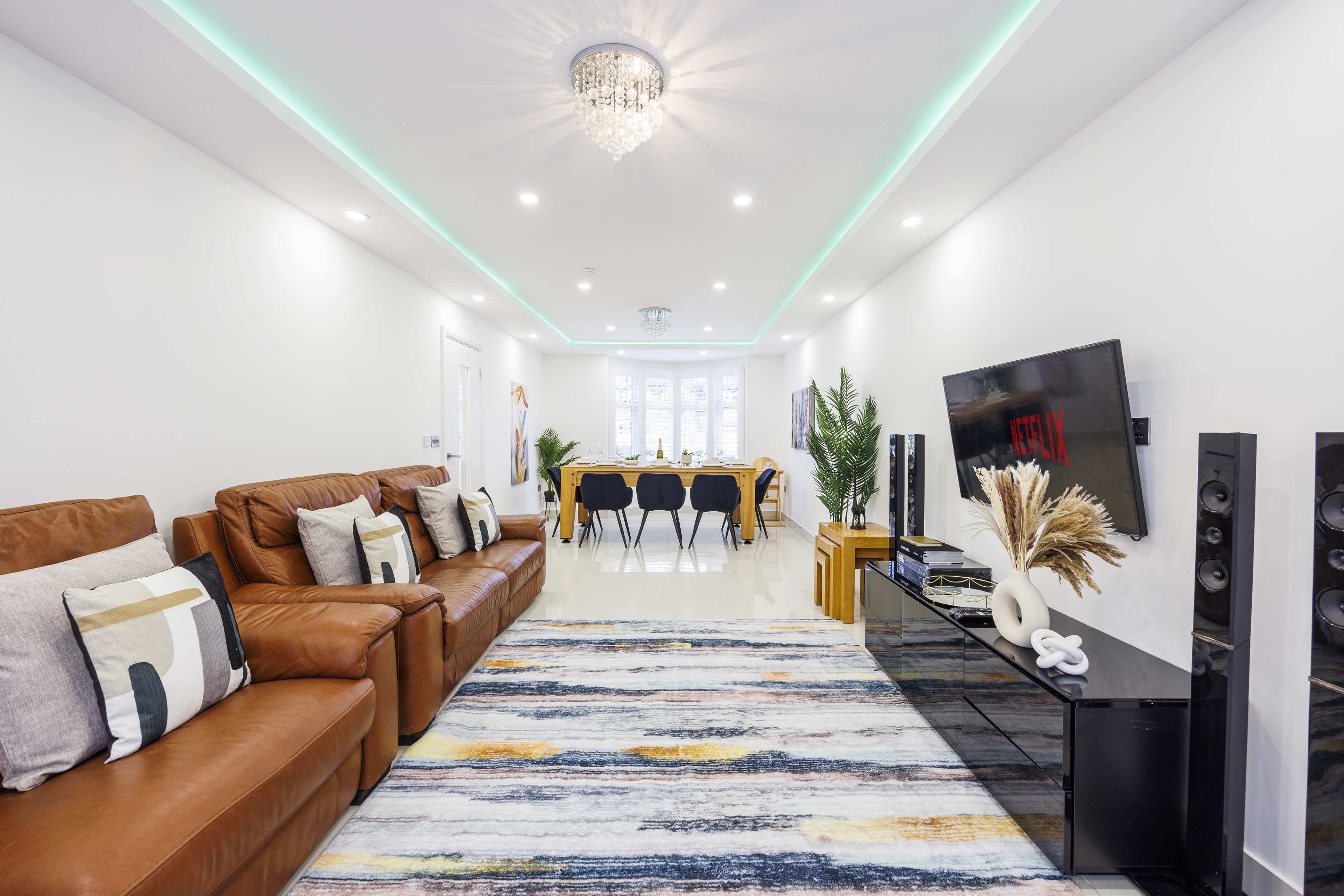 A modern living room features a brown leather sofa, a patterned rug, and a sleek entertainment center with a large TV, leading into a dining area with black chairs.