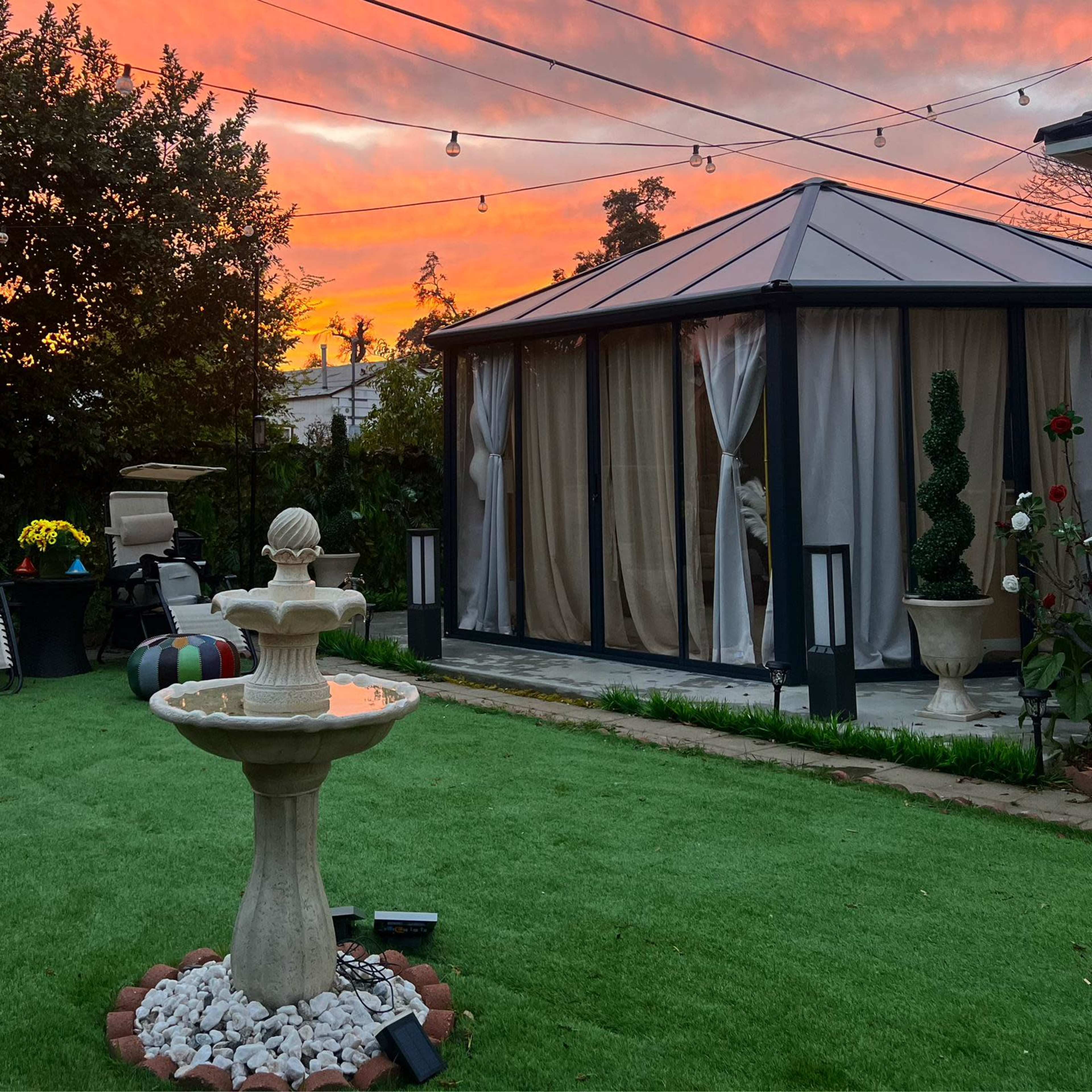 A fountain stands in the foreground of a backyard, with a gazebo in the background illuminated by a vibrant sunset.