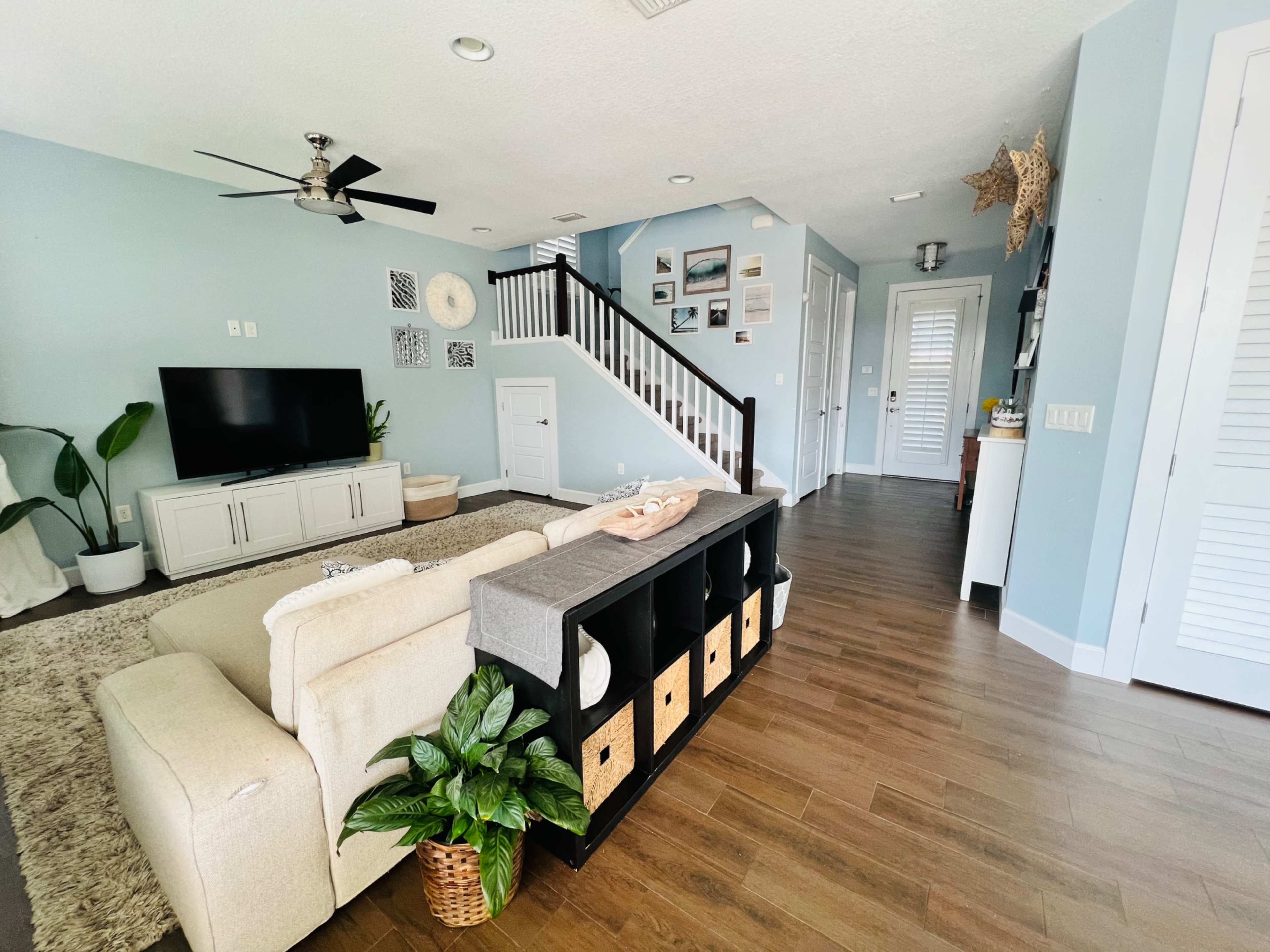 The image shows a modern living room with a light-colored sofa, a television mounted on the wall, and a staircase leading to an upper level, all set against a pale blue wall.
