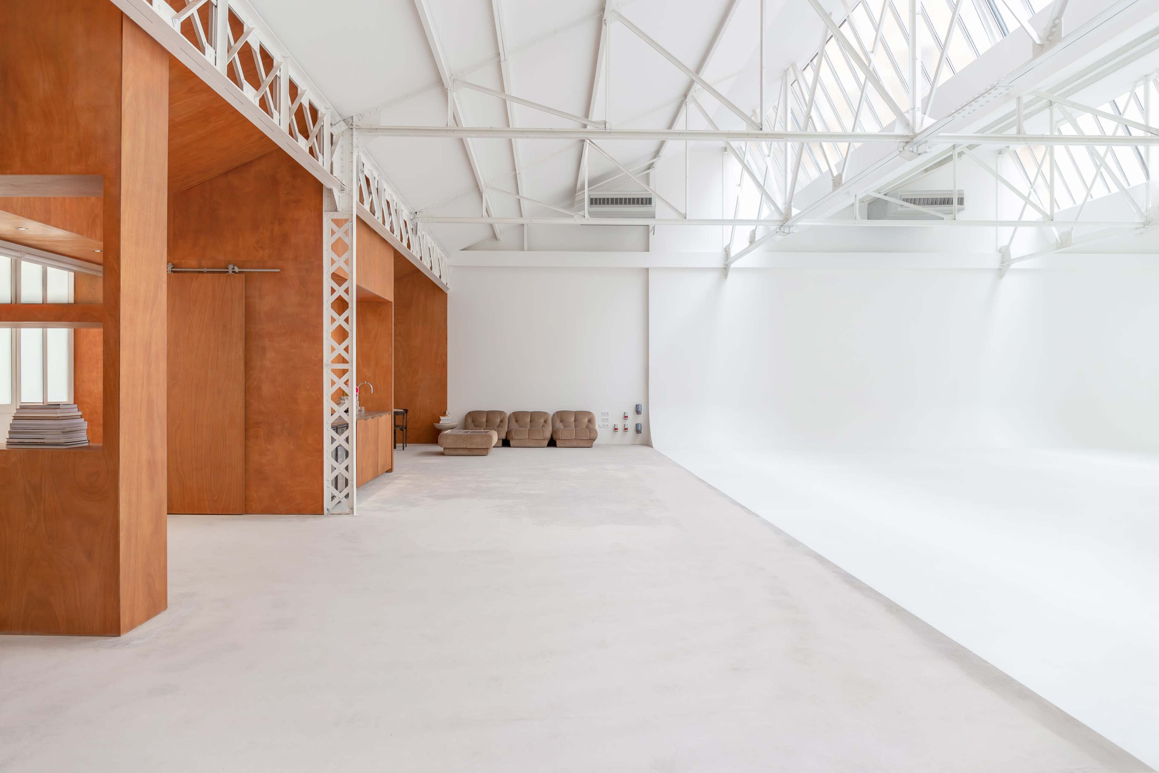 The image shows a spacious, minimalistic interior with light-colored walls, wooden accents, and a few brown couches in a well-lit area.