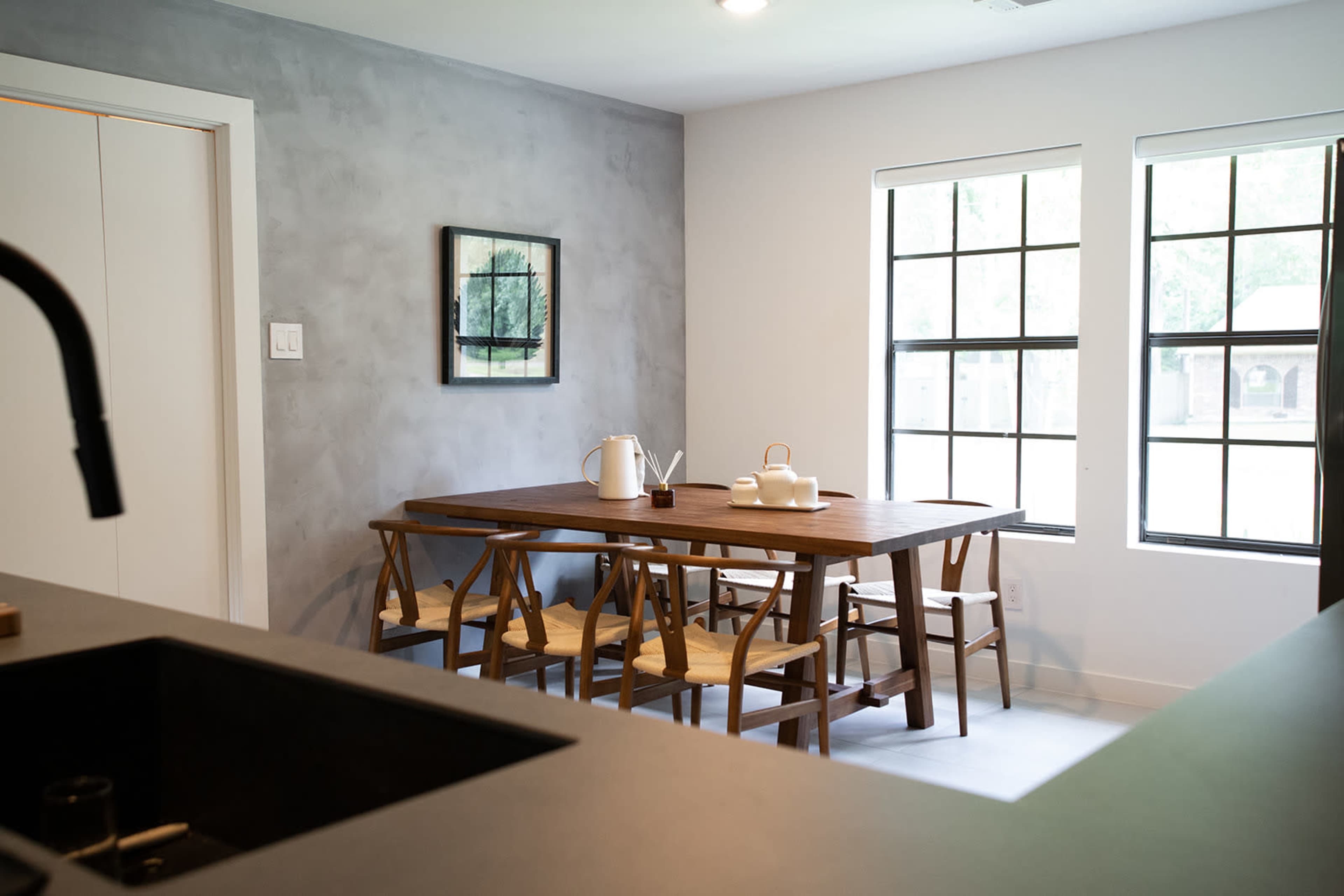 A modern dining area features a wooden table surrounded by chairs, with a minimalistic decor against a gray wall and large windows providing natural light.