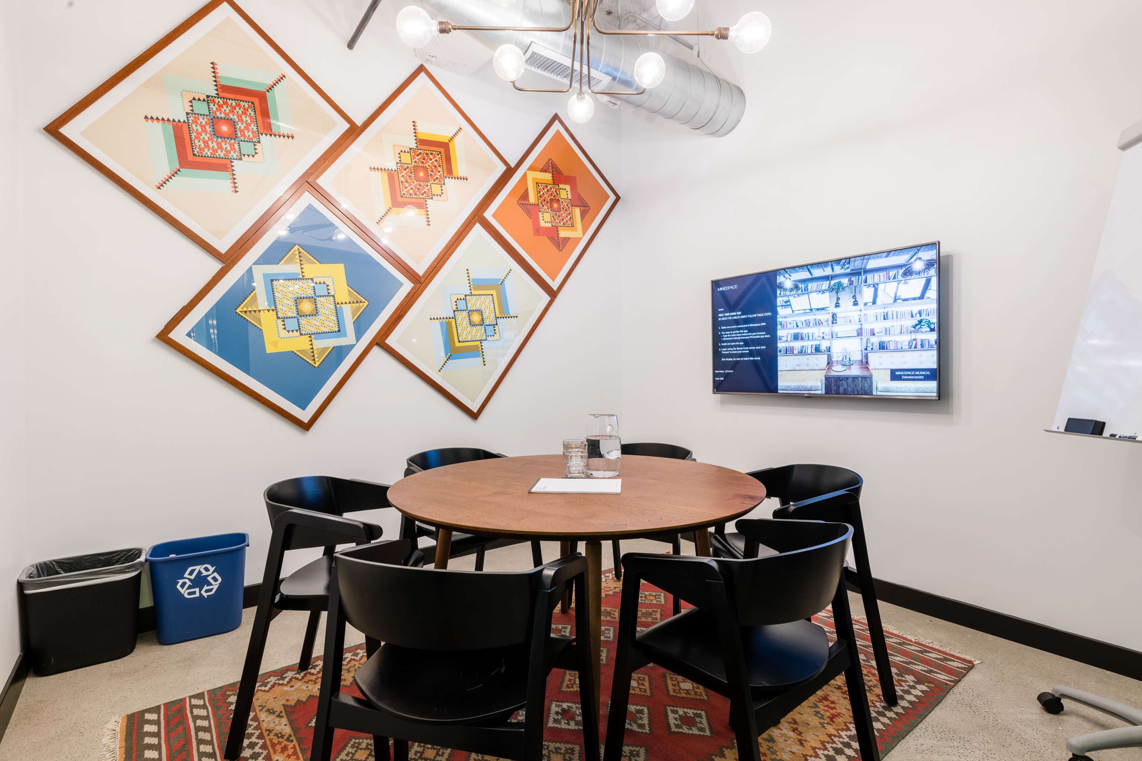 A conference room features a round wooden table surrounded by black chairs, with colorful wall art and a television displaying information.