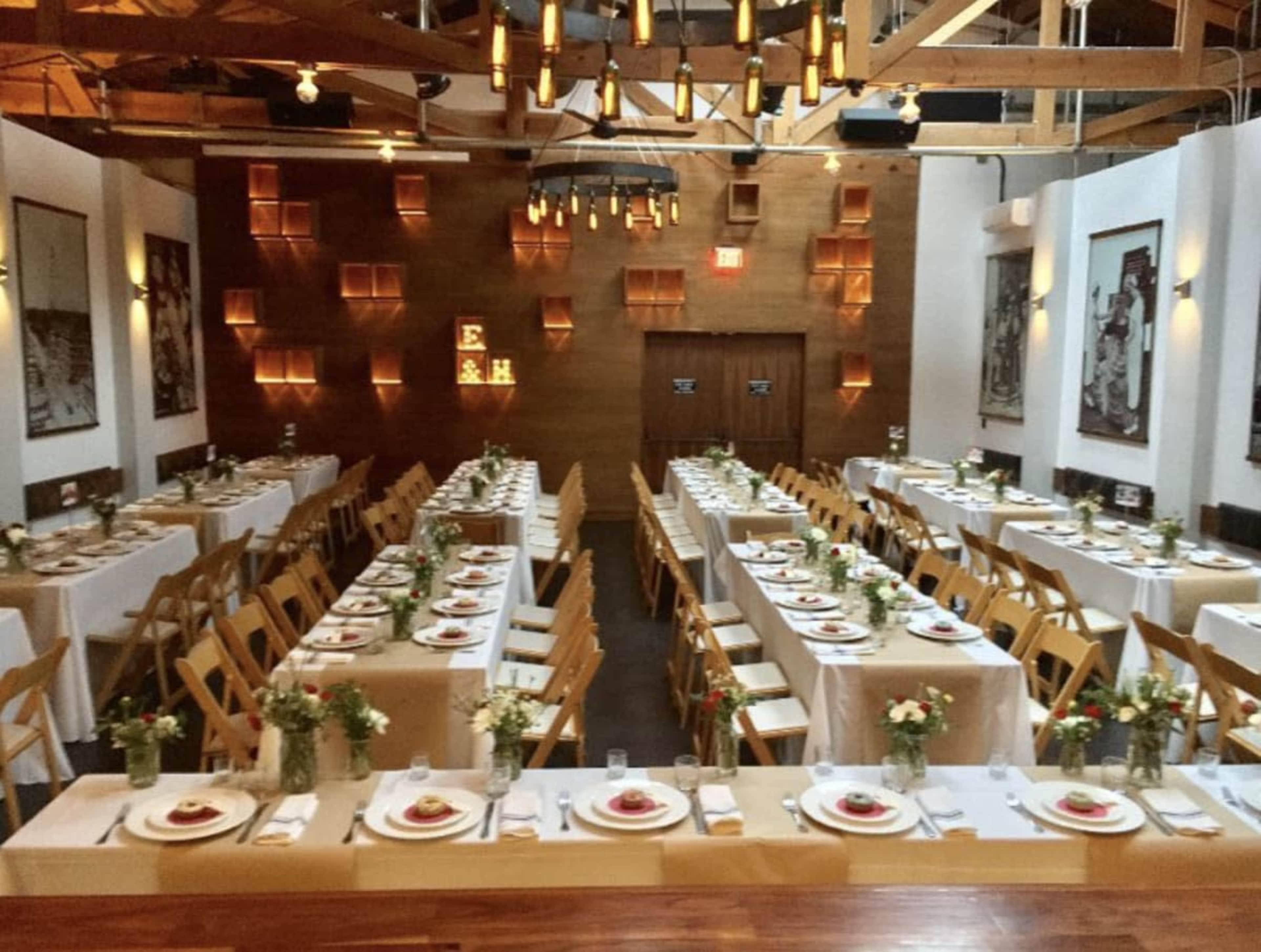 A spacious dining area is arranged with long tables set for a formal event, featuring centerpieces of flowers and neatly arranged plates.