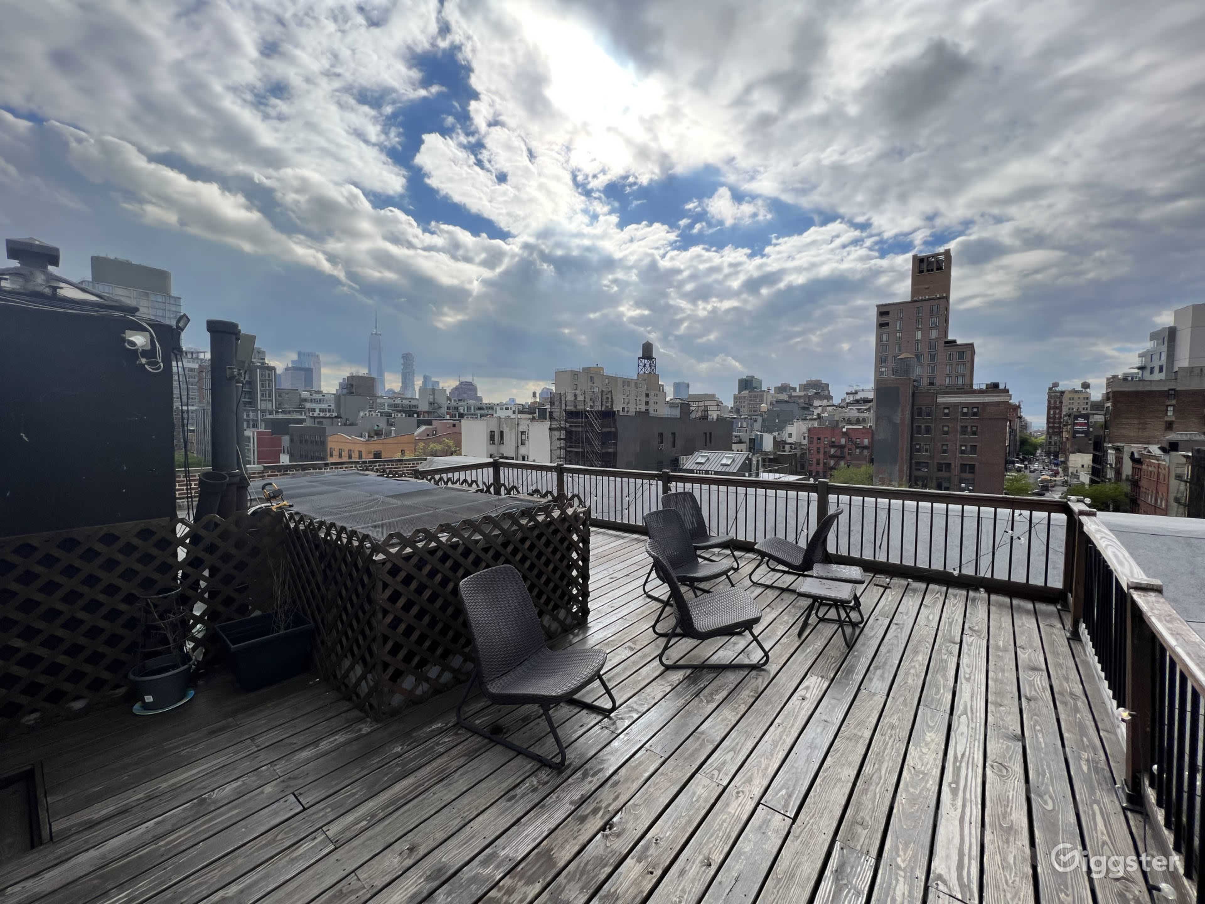 A rooftop terrace with wooden decking, several chairs, and a view of a city skyline under a cloudy sky.
