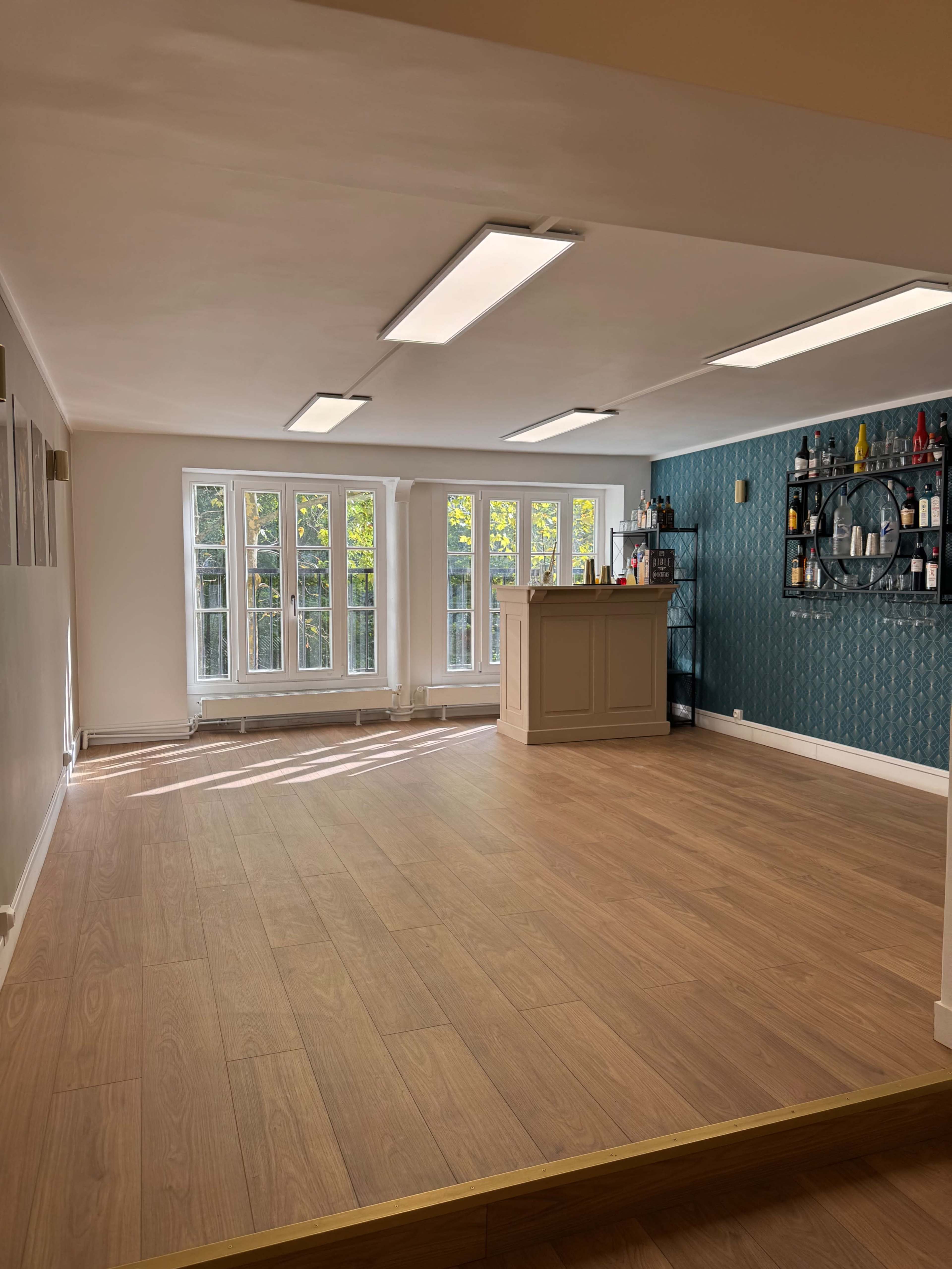The image shows an empty interior space with wood flooring, a bar counter against a patterned wall, and large windows allowing natural light to enter.