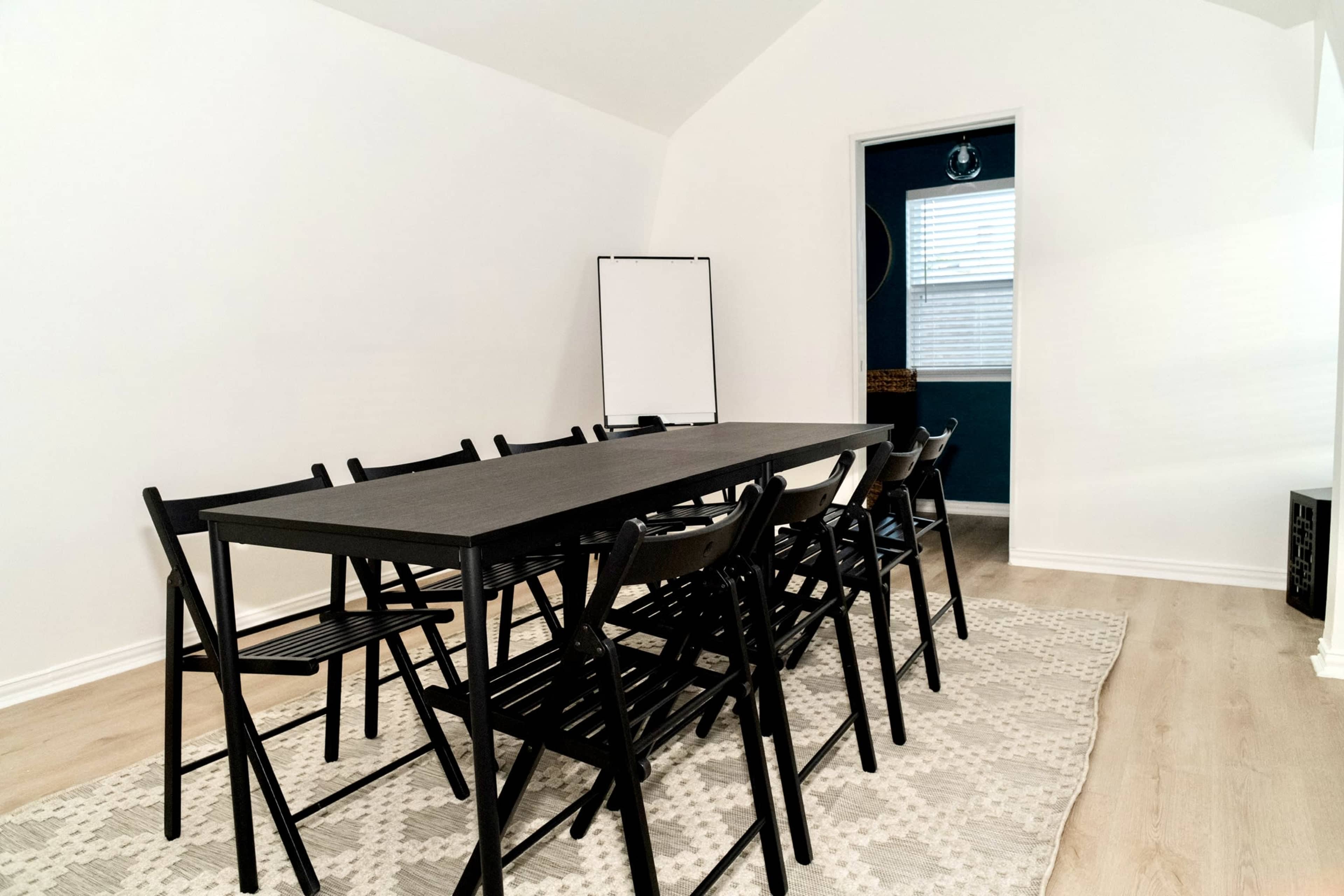 A long black table surrounded by eight folding chairs is set in a light-colored room with an open door leading to a blue-walled space.