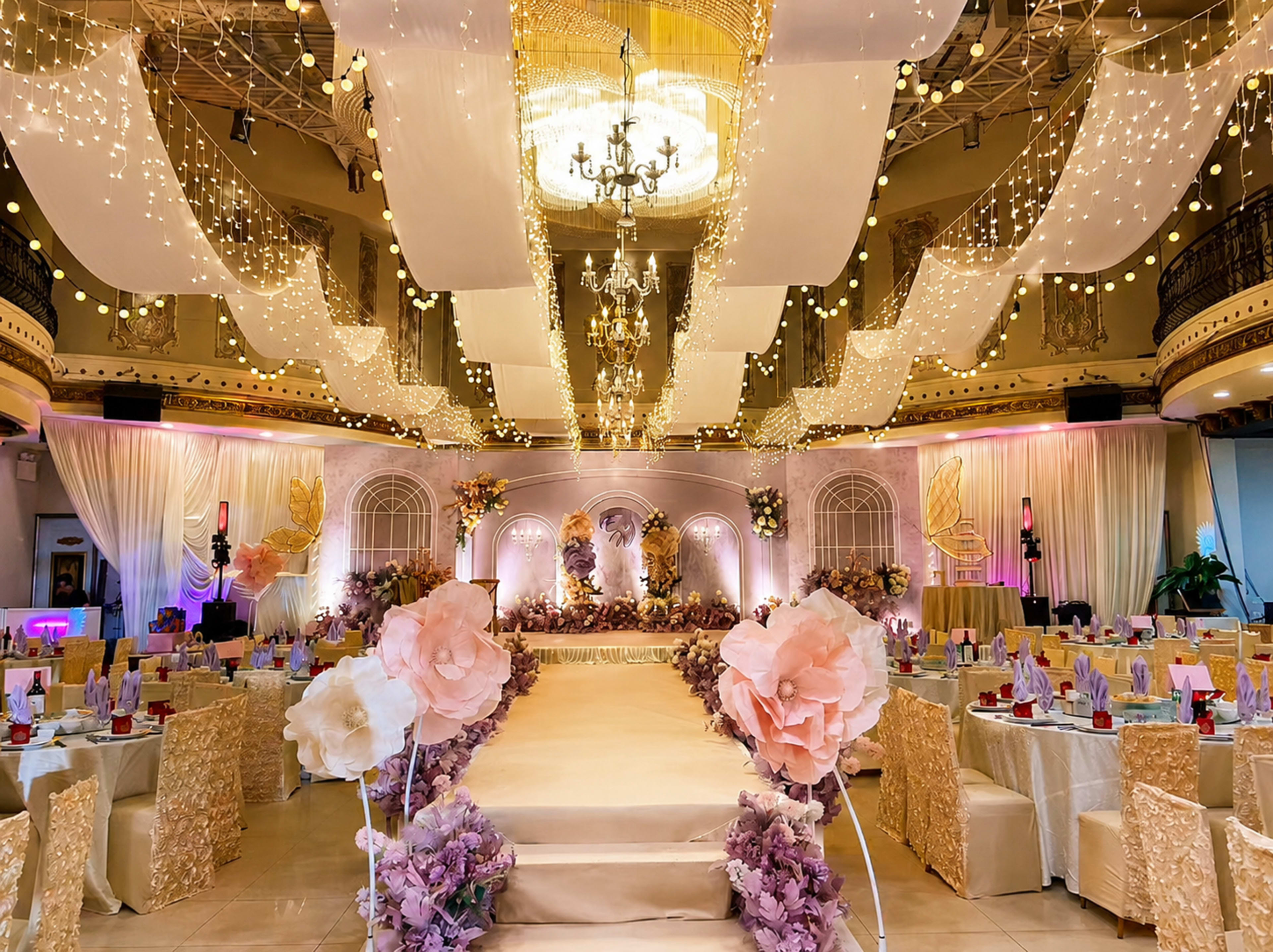 The image shows a beautifully decorated banquet hall set up for a formal event, featuring elegant table settings, draped fabric, fairy lights, and a central stage with floral arrangements.