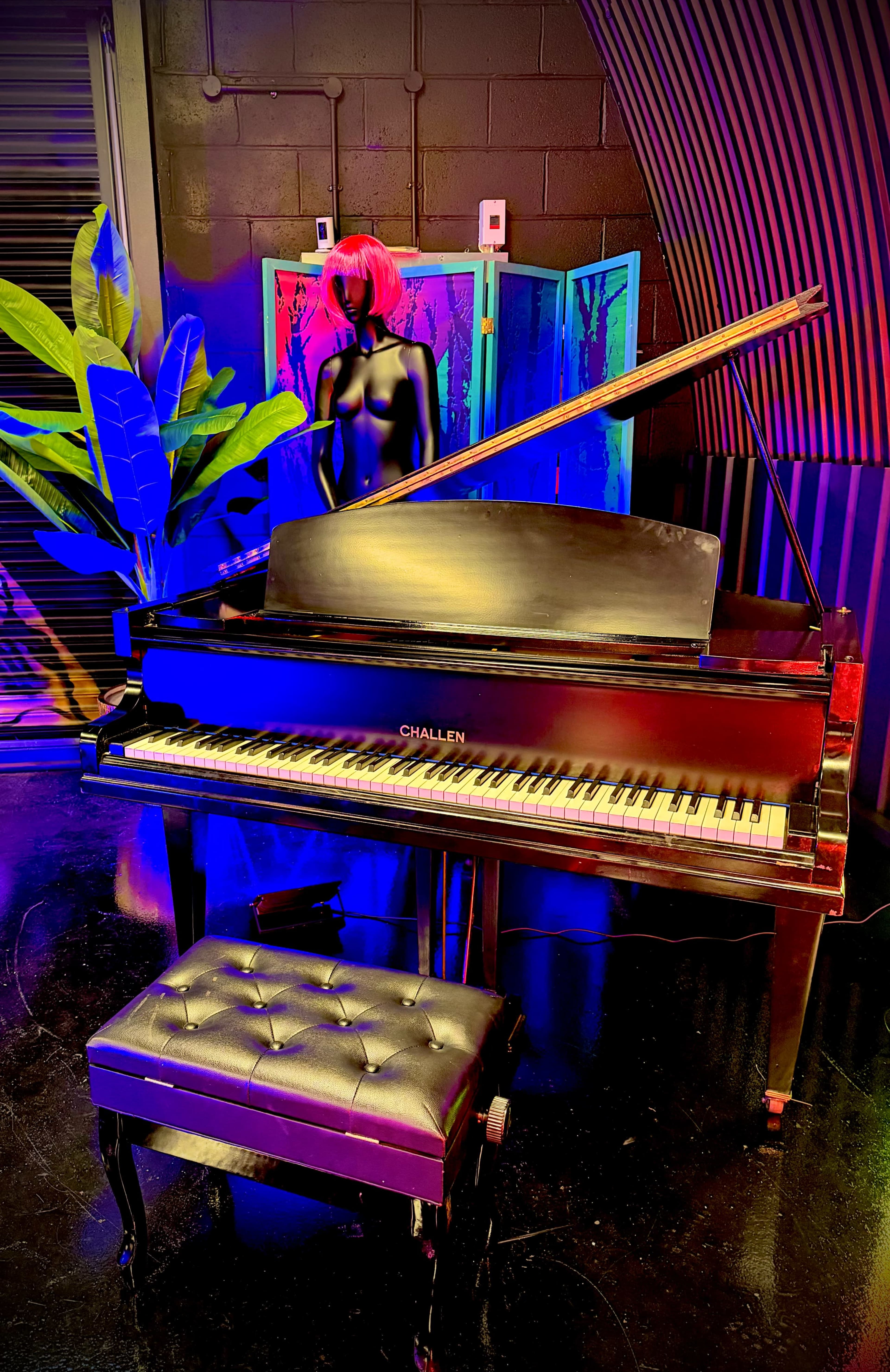 A grand piano is positioned in front of a colorful backdrop, accompanied by a black mannequin and a large plant.
