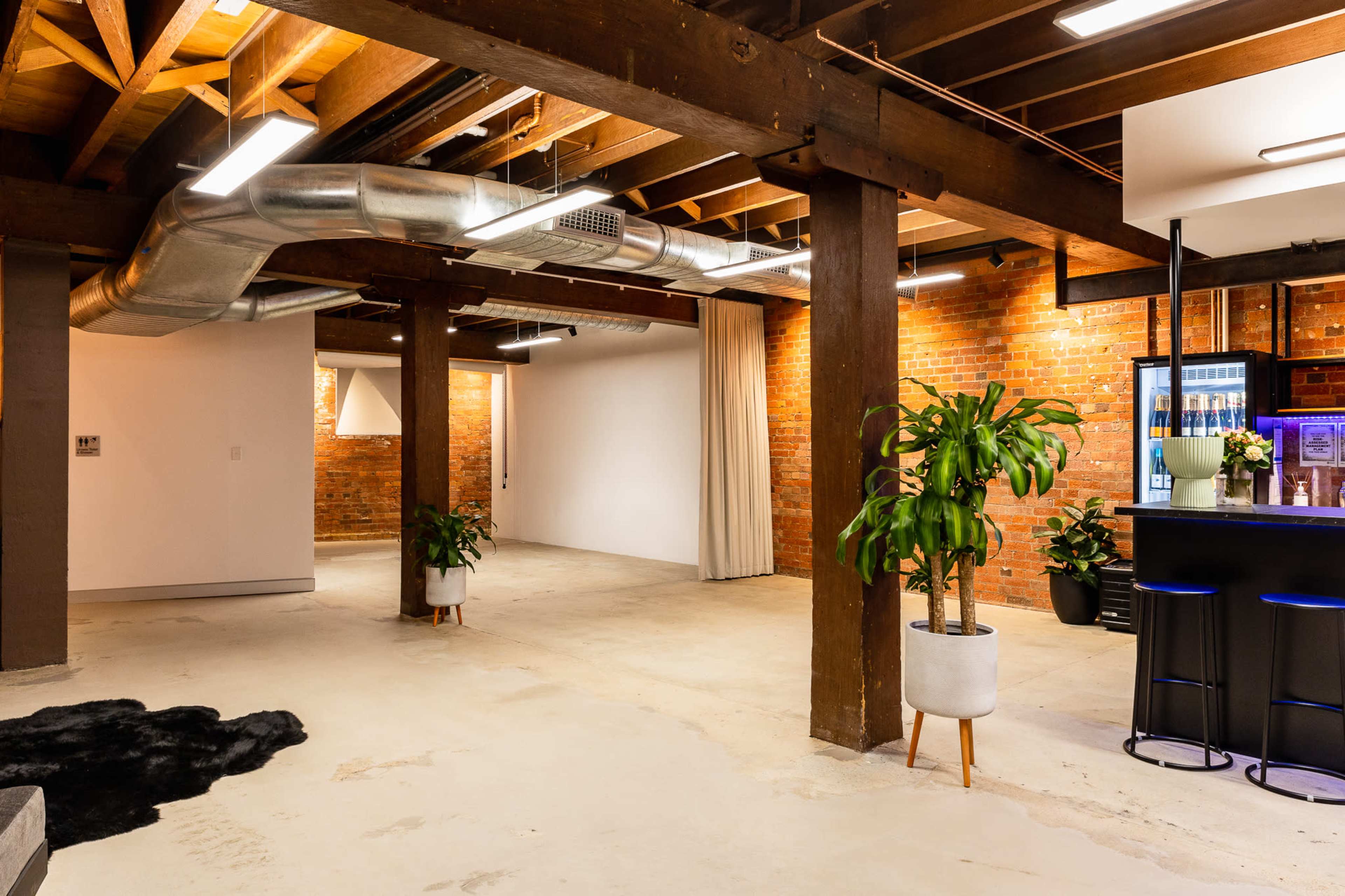 An industrial-style interior with exposed brick walls, wooden beams, polished concrete floors, and a bar area at the end.