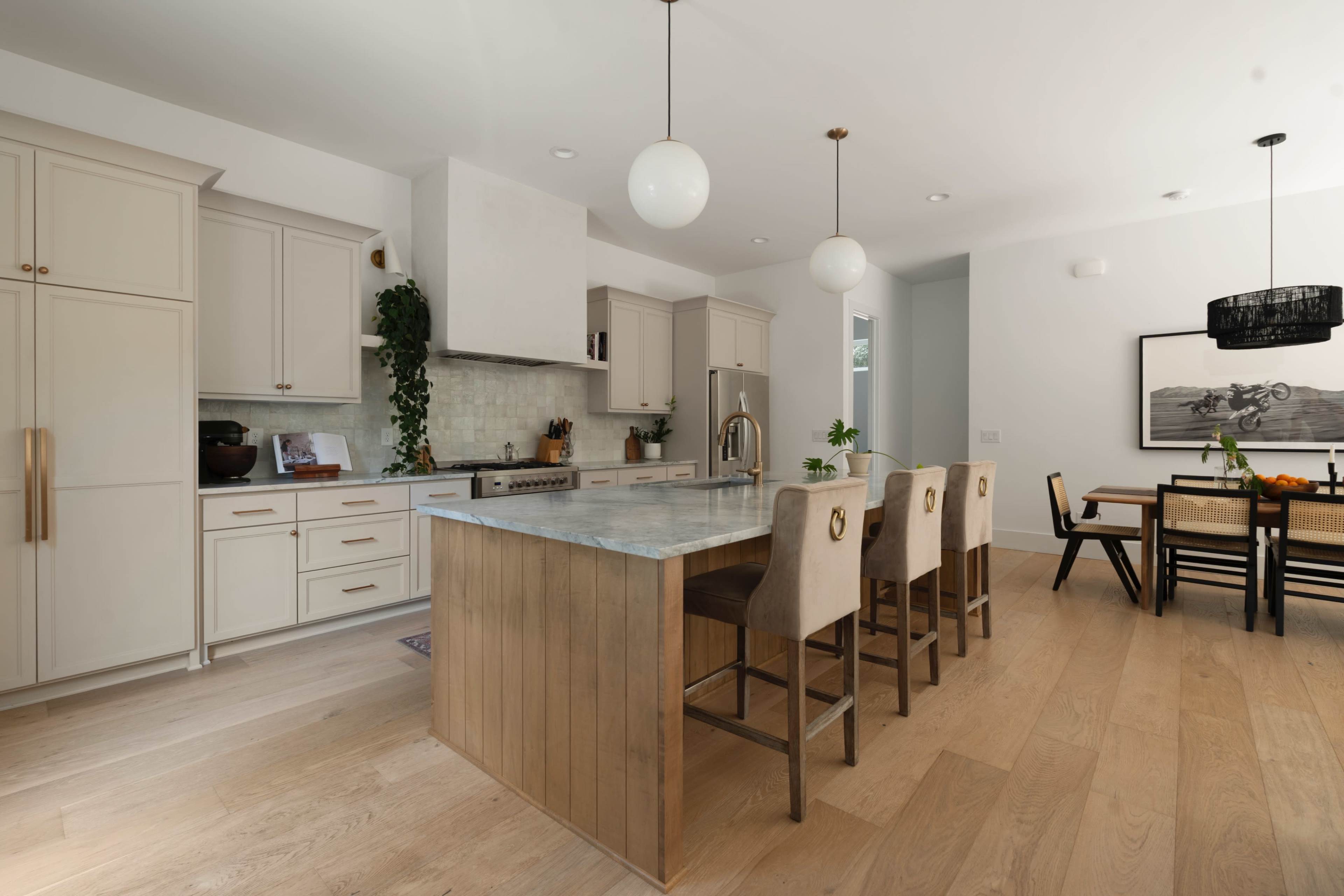 A modern kitchen features light cabinetry, a large island with seating, and an adjacent dining area with a wooden table.