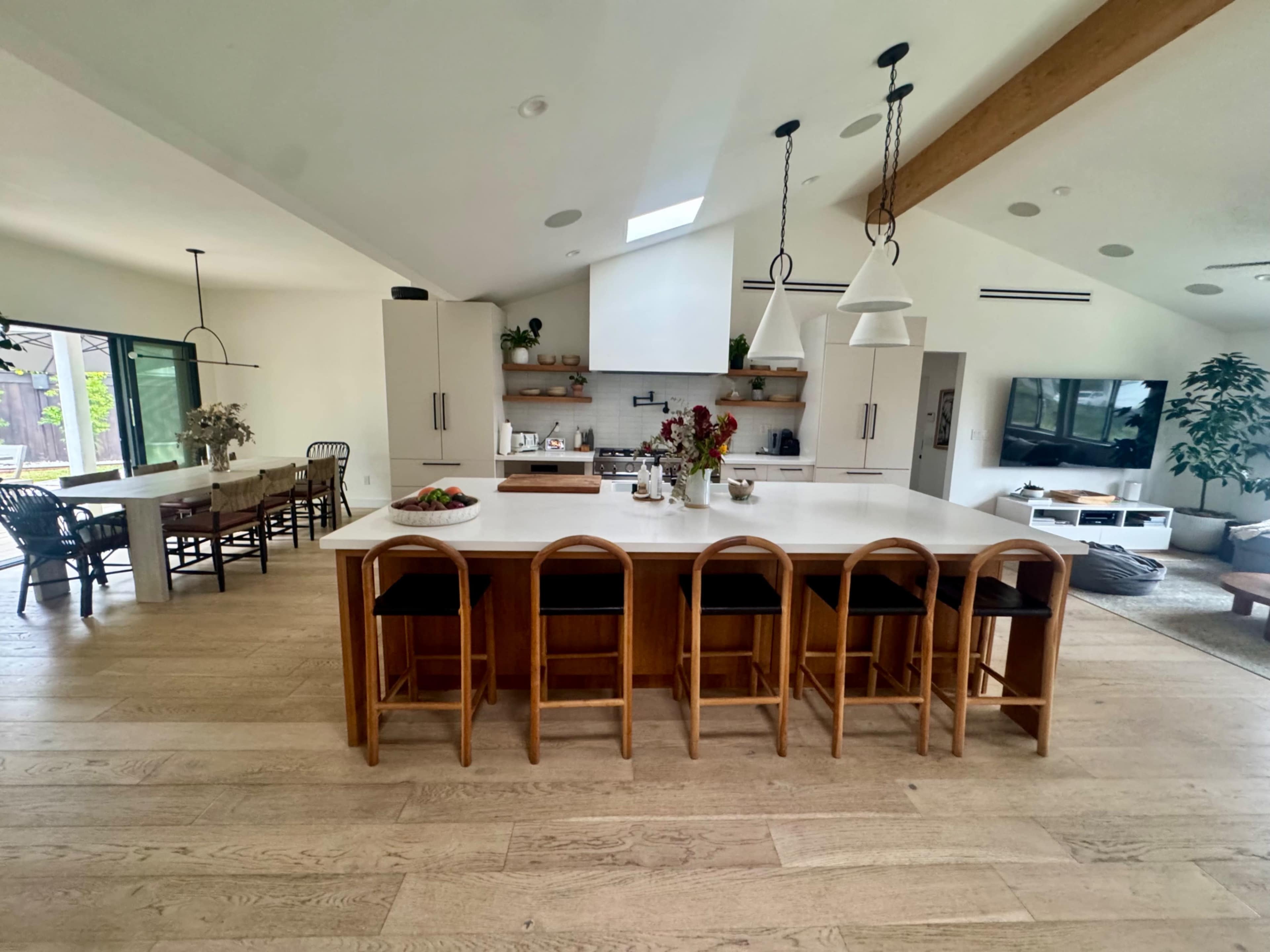 The image shows a modern kitchen with a large island, wooden bar stools, and an open dining area featuring a wooden table and chairs.