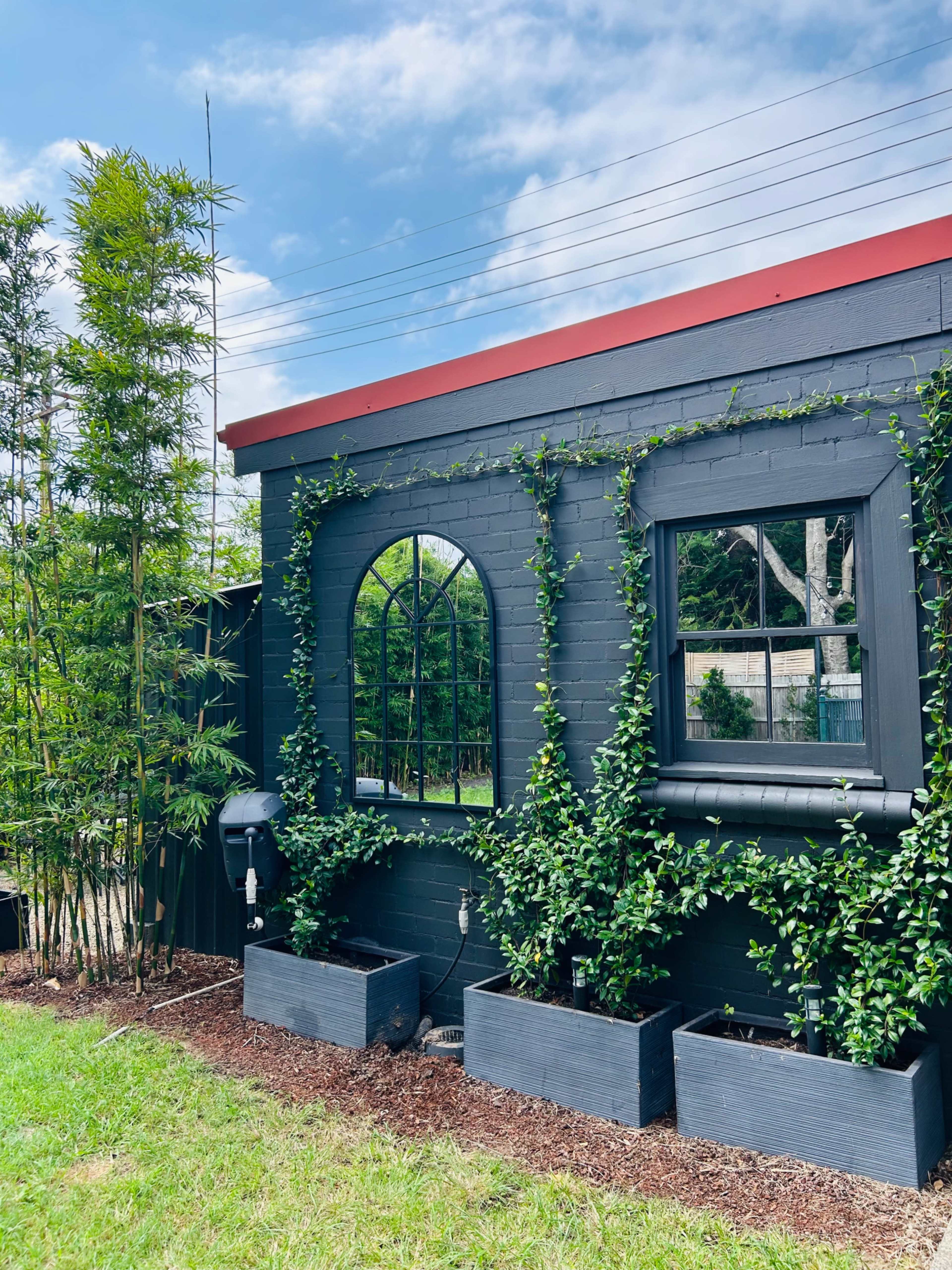 A black-painted building exterior with a red roof, featuring two arched mirrors and green climbing plants along the walls, surrounded by rectangular planters filled with vegetation.
